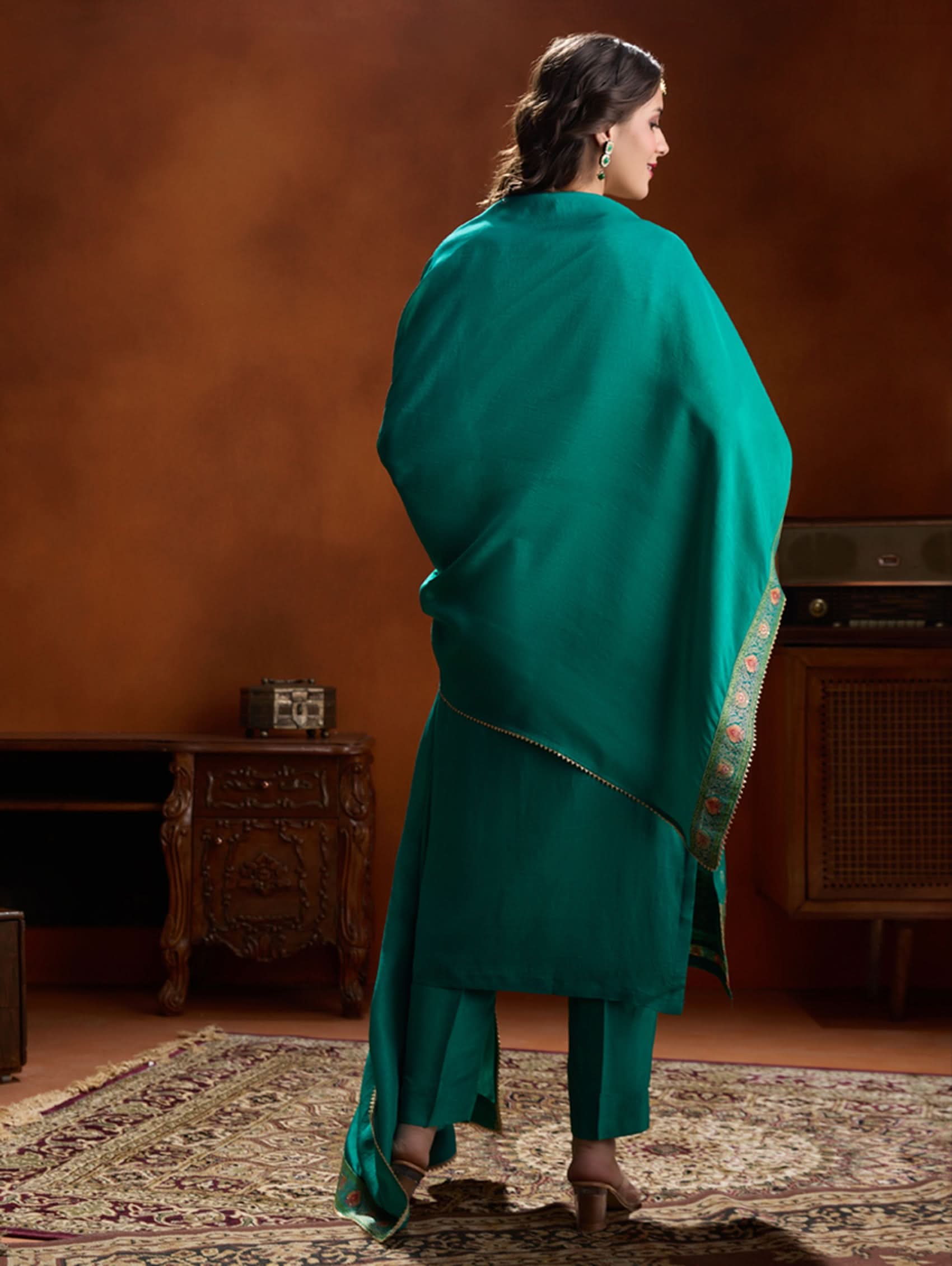 Woman in a teal traditional outfit standing in a room with wooden furniture and a patterned rug.