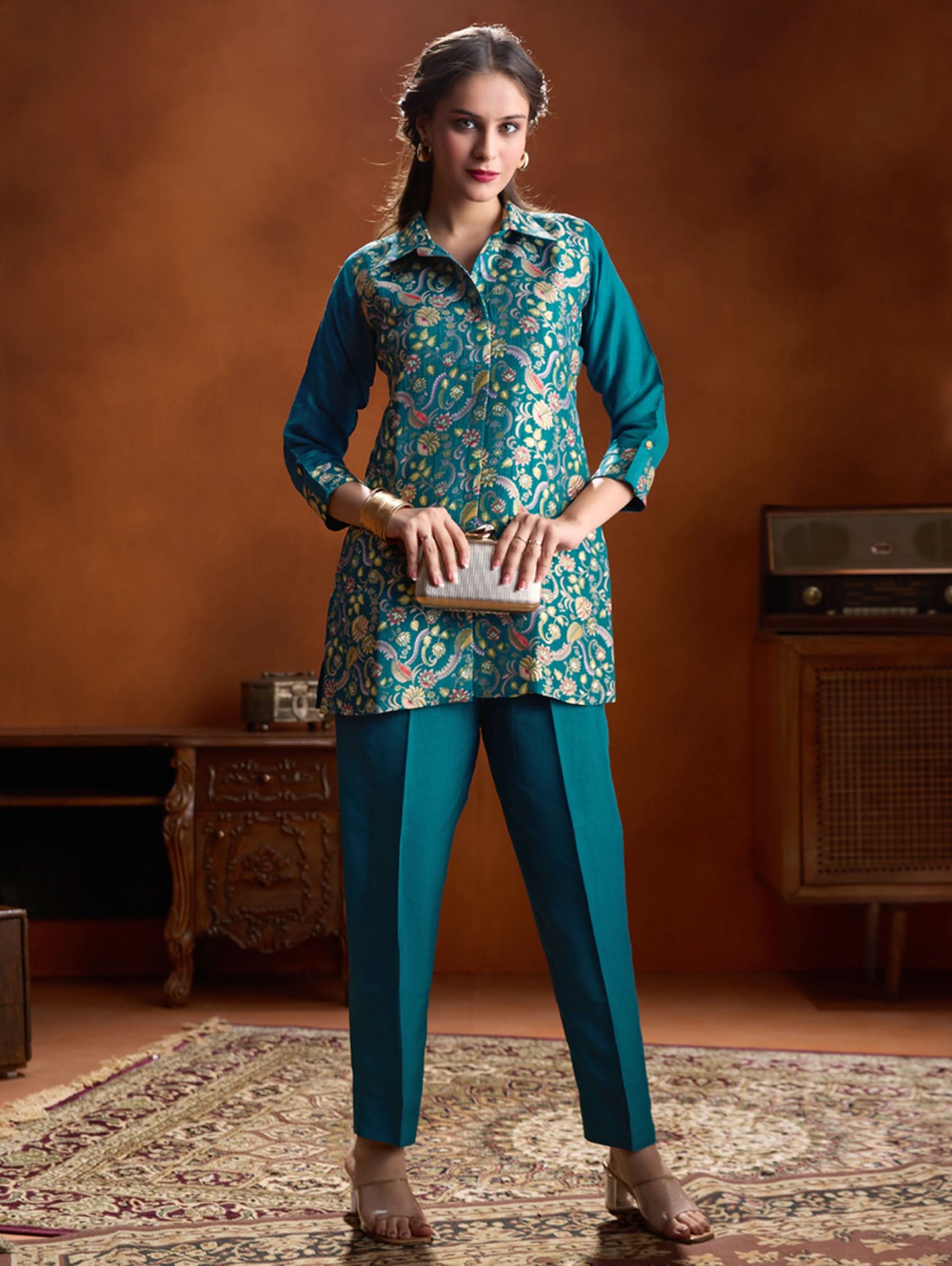 Woman in a teal and floral outfit standing in a room with a warm brown wall.