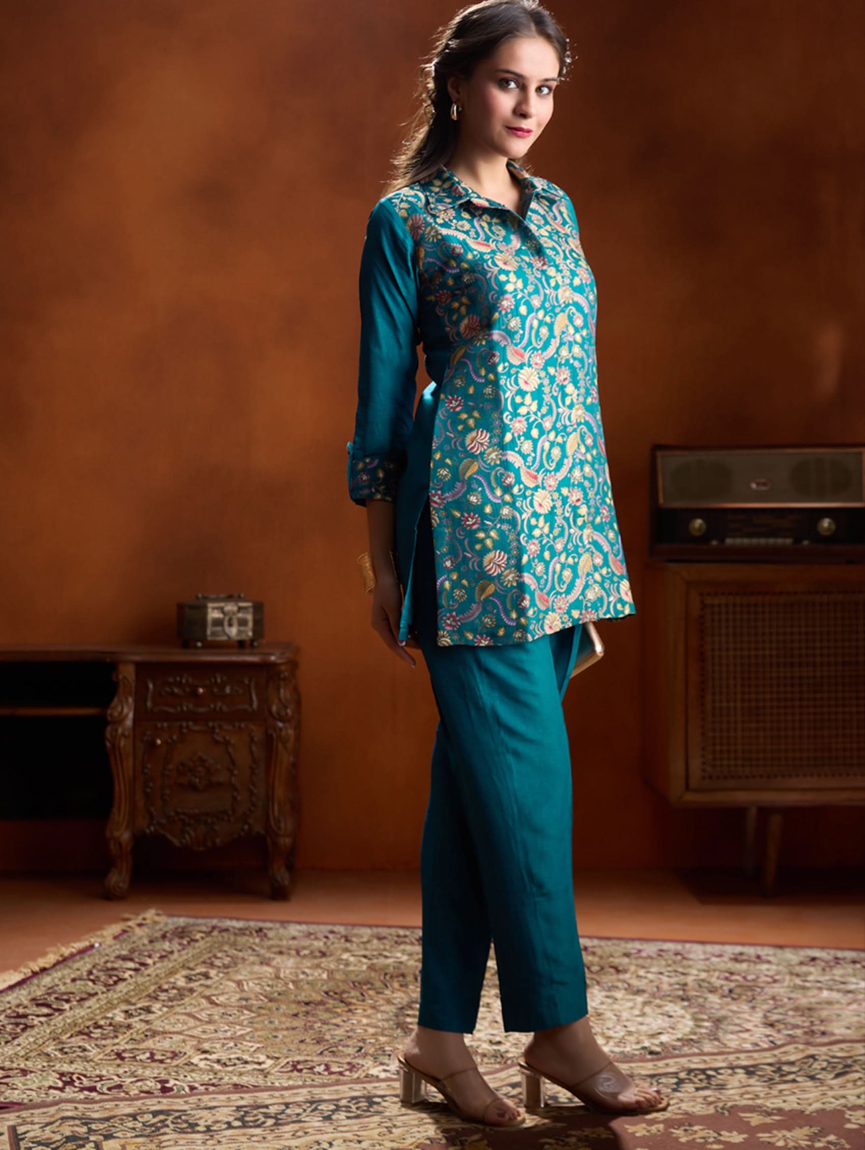 Woman wearing a teal floral top and pants in a room with vintage decor.