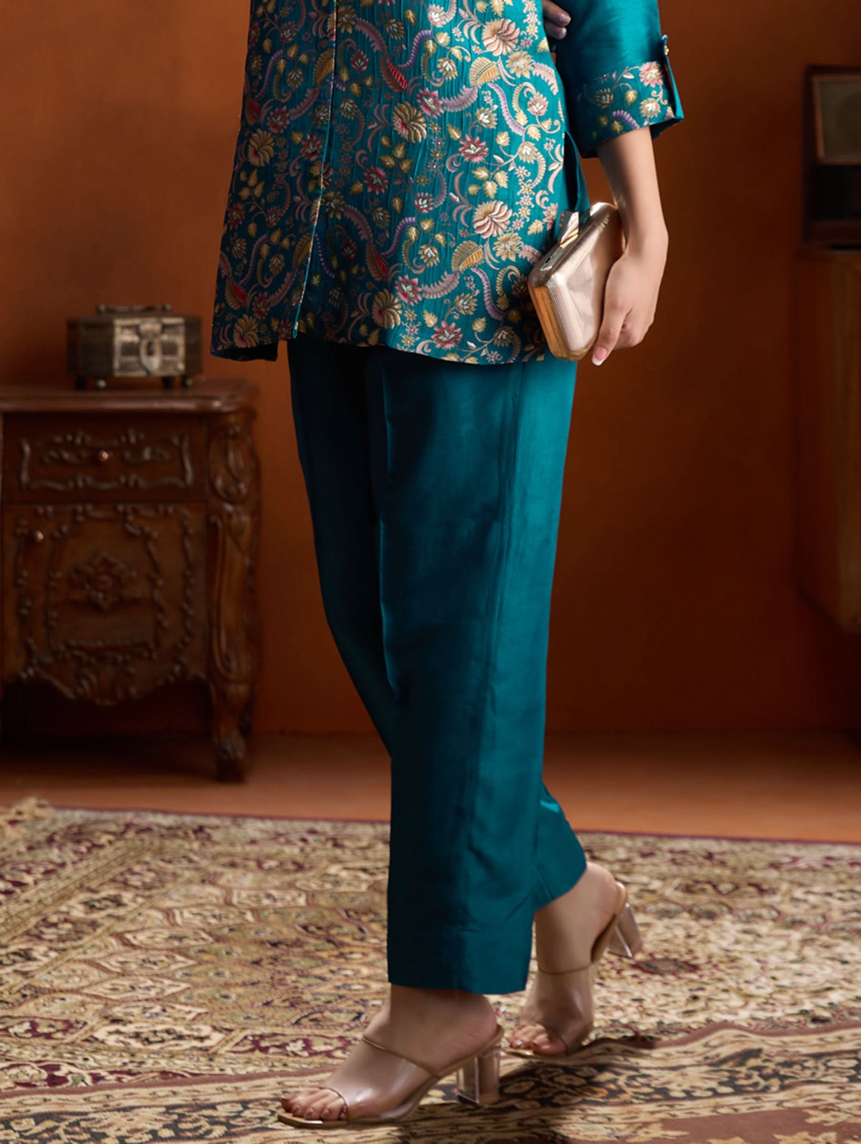 Person wearing a teal embroidered top and pants with a gold clutch against an orange wall.