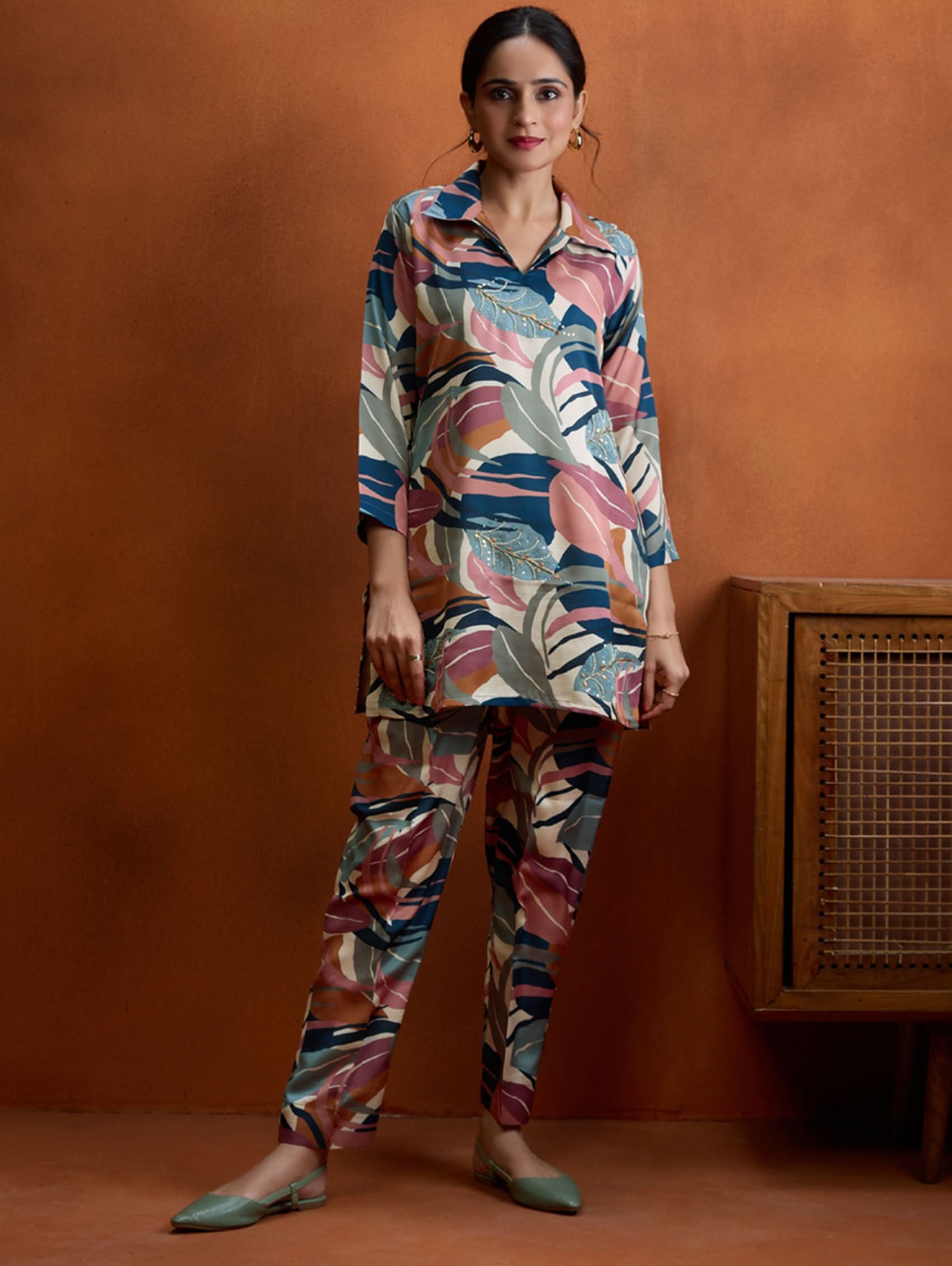 A woman standing against an orange background, wearing a digitally printed satin co-ord set with a short kurta and pants, featuring a revere collar and hand embroidery on the kurta.