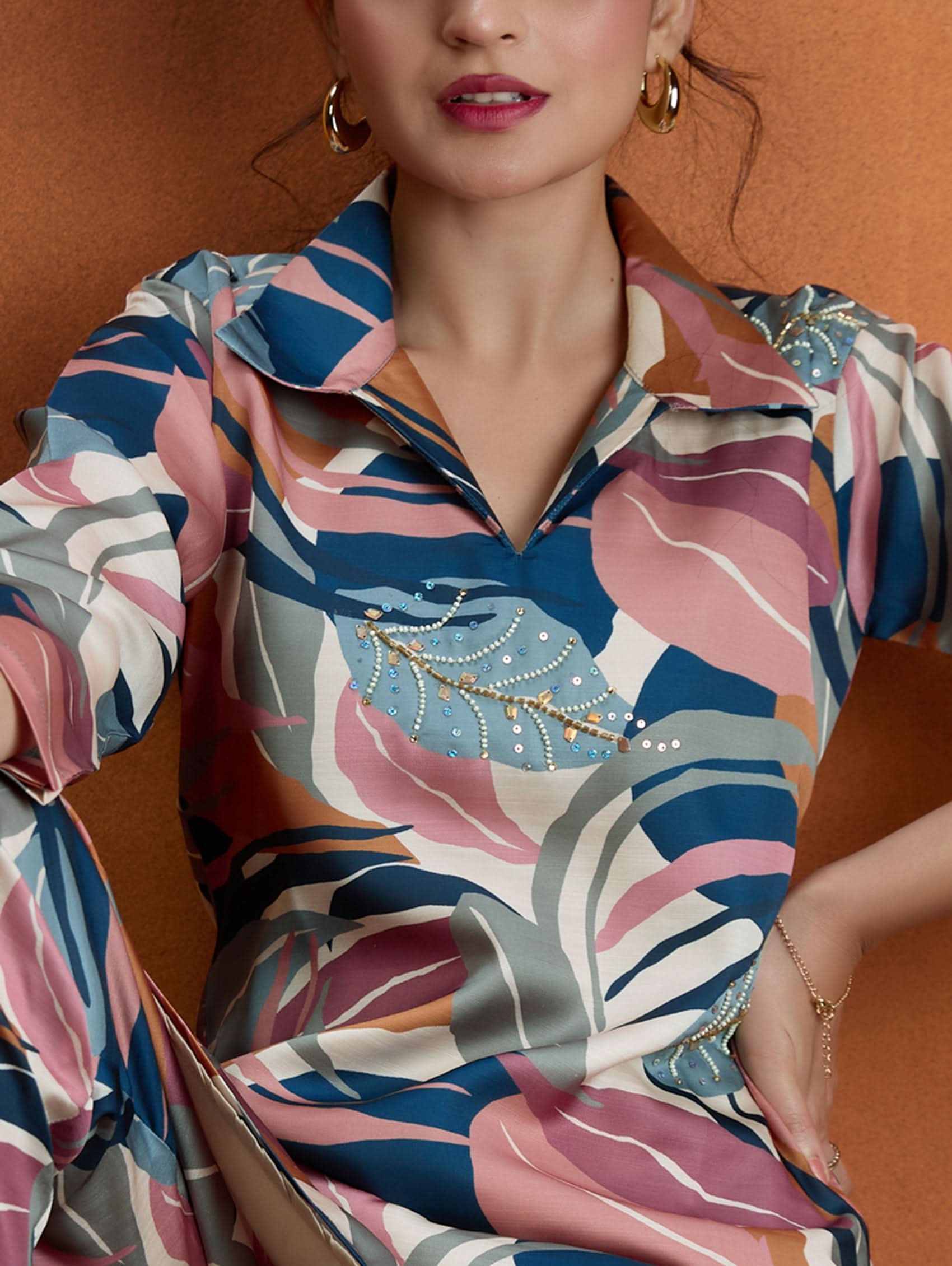 A woman standing against an orange background, wearing a digitally printed satin co-ord set with a short kurta and pants, featuring a revere collar and hand embroidery on the kurta.