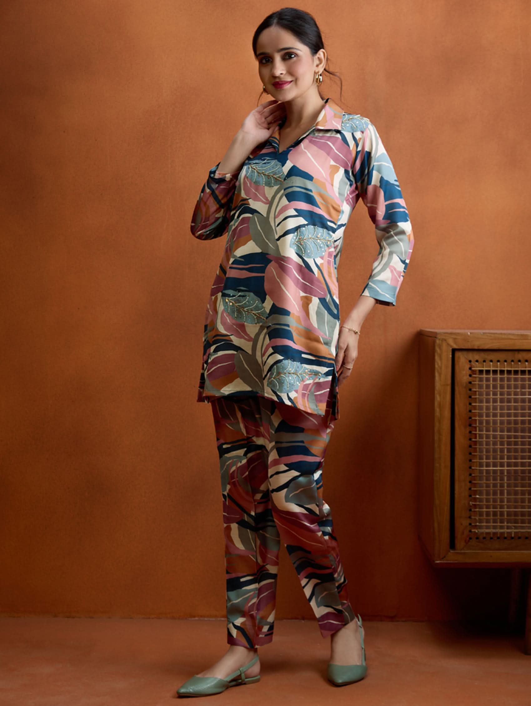 A woman standing against an orange background, wearing a digitally printed satin co-ord set with a short kurta and pants, featuring a revere collar and hand embroidery on the kurta.