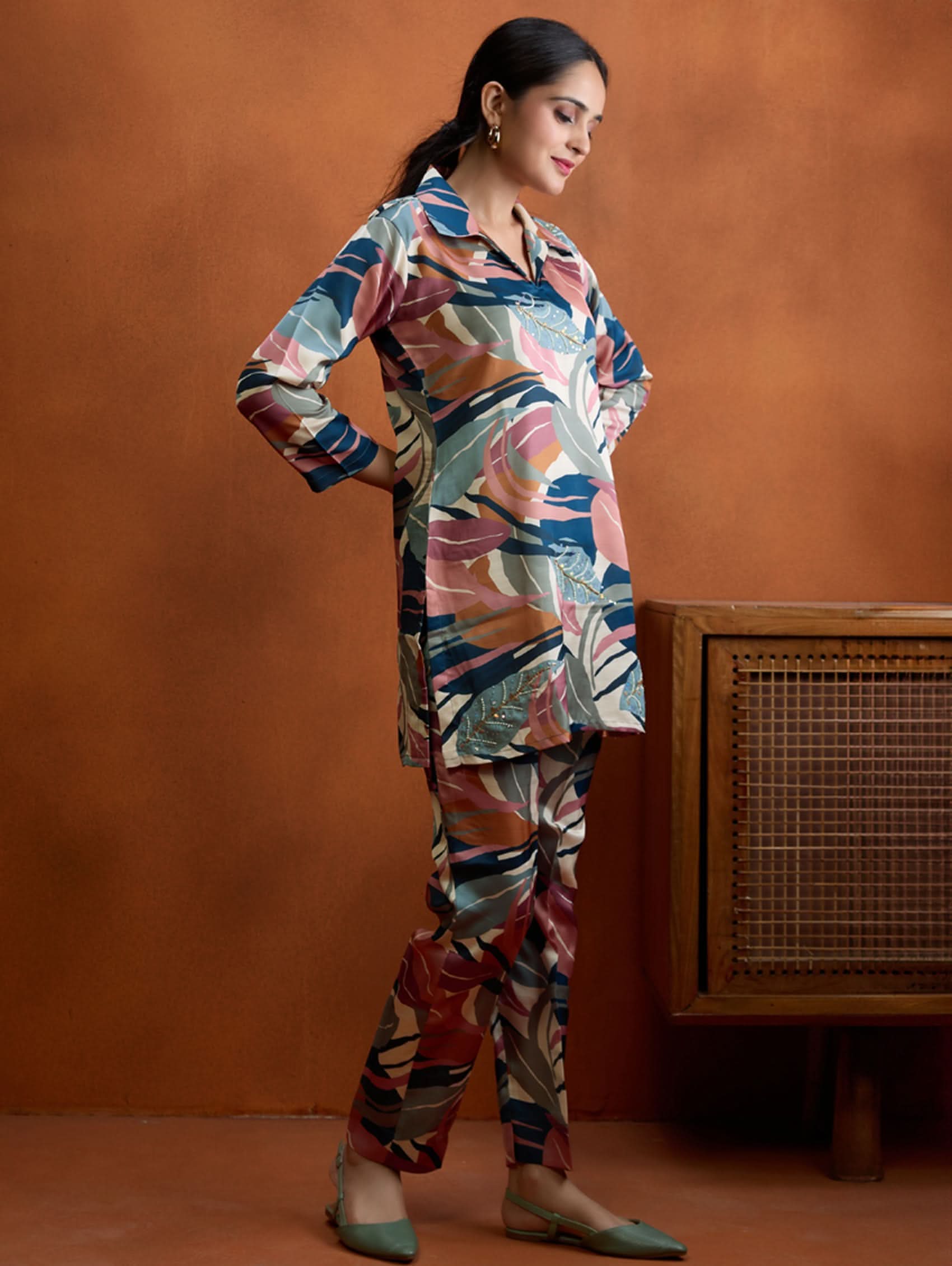 A woman standing against an orange background, wearing a digitally printed satin co-ord set with a short kurta and pants, featuring a revere collar and hand embroidery on the kurta.