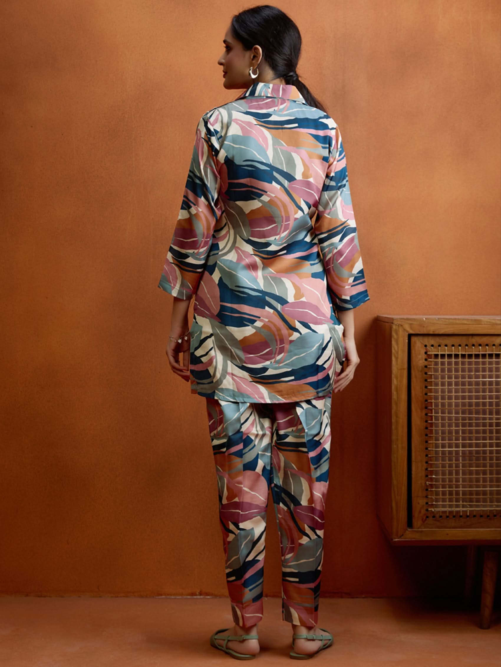 A woman standing against an orange background, wearing a digitally printed satin co-ord set with a short kurta and pants, featuring a revere collar and hand embroidery on the kurta.