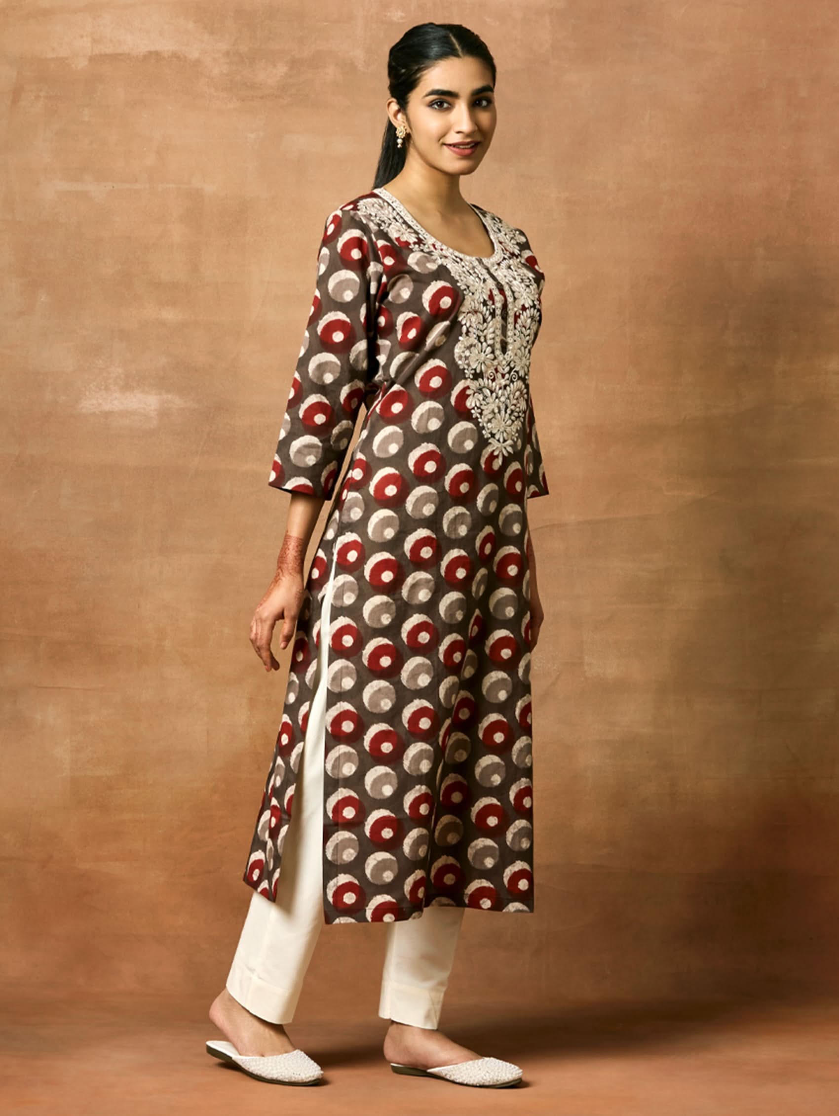 Woman wearing a patterned kurta and white pants against a brown background