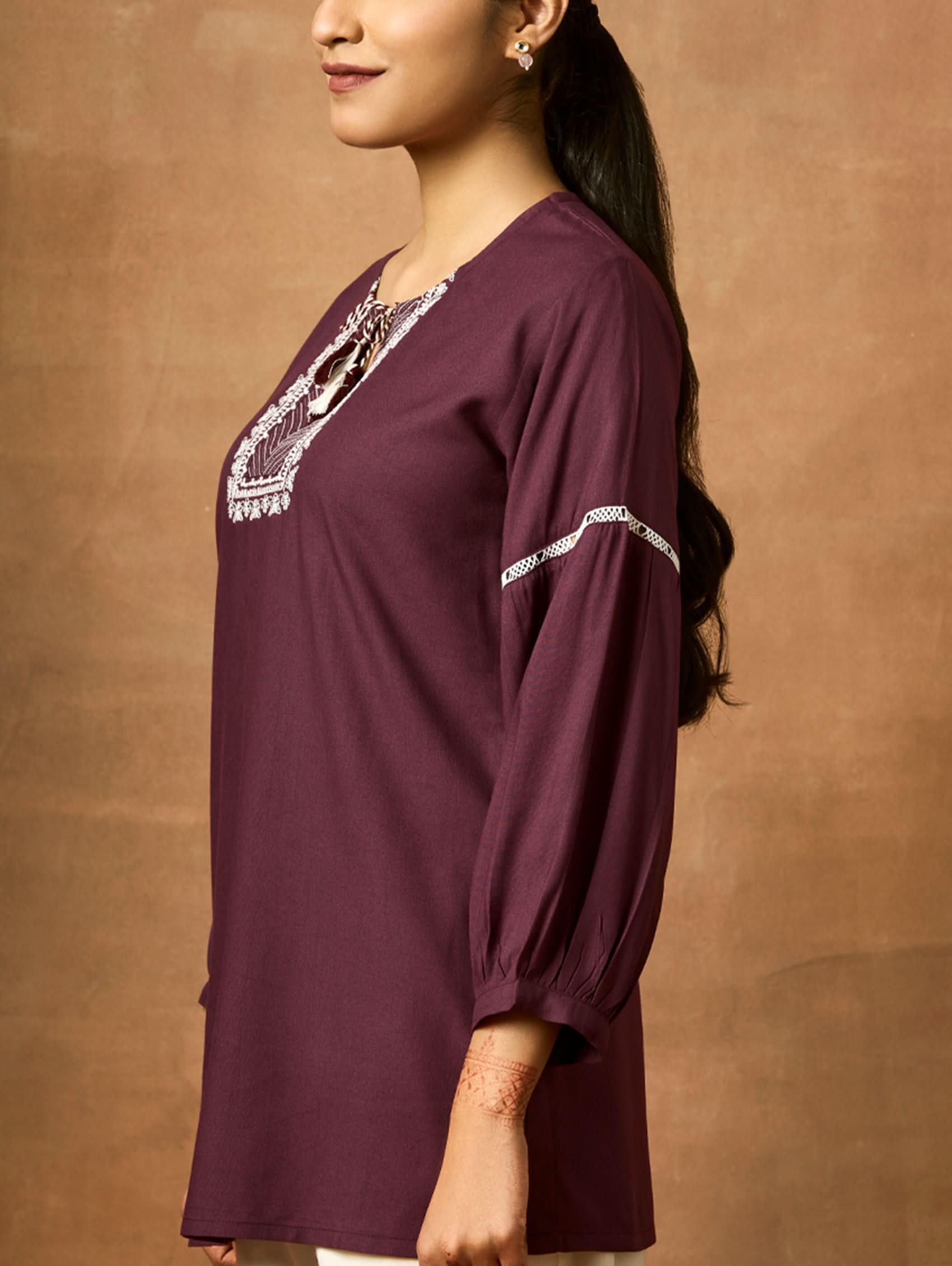 Woman wearing a purple long-sleeve top with white embroidery against a brown background.