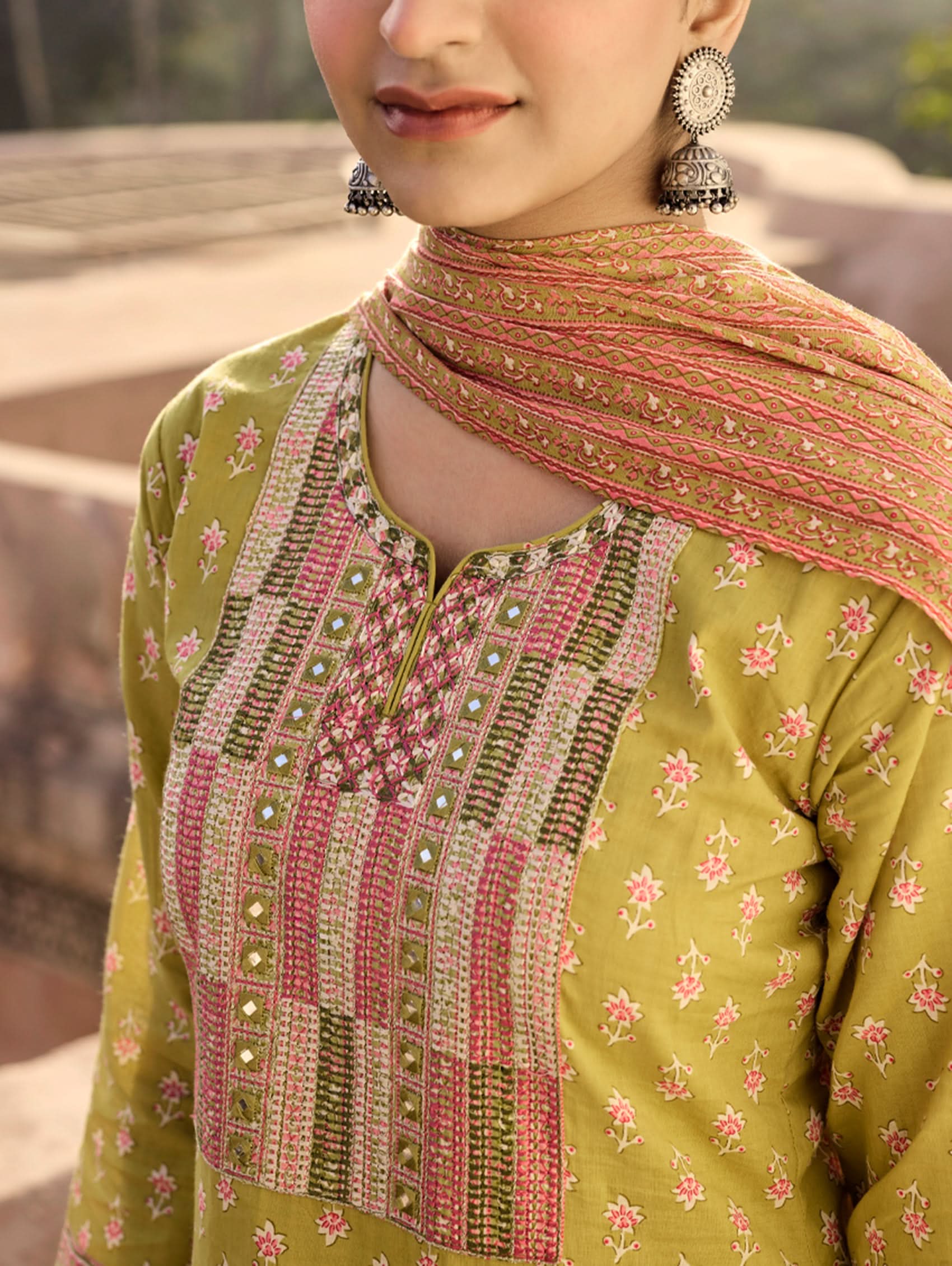 A woman wearing a green and orange cotton suit set with embroidery, consisting of a kurta with pants and a dupatta.