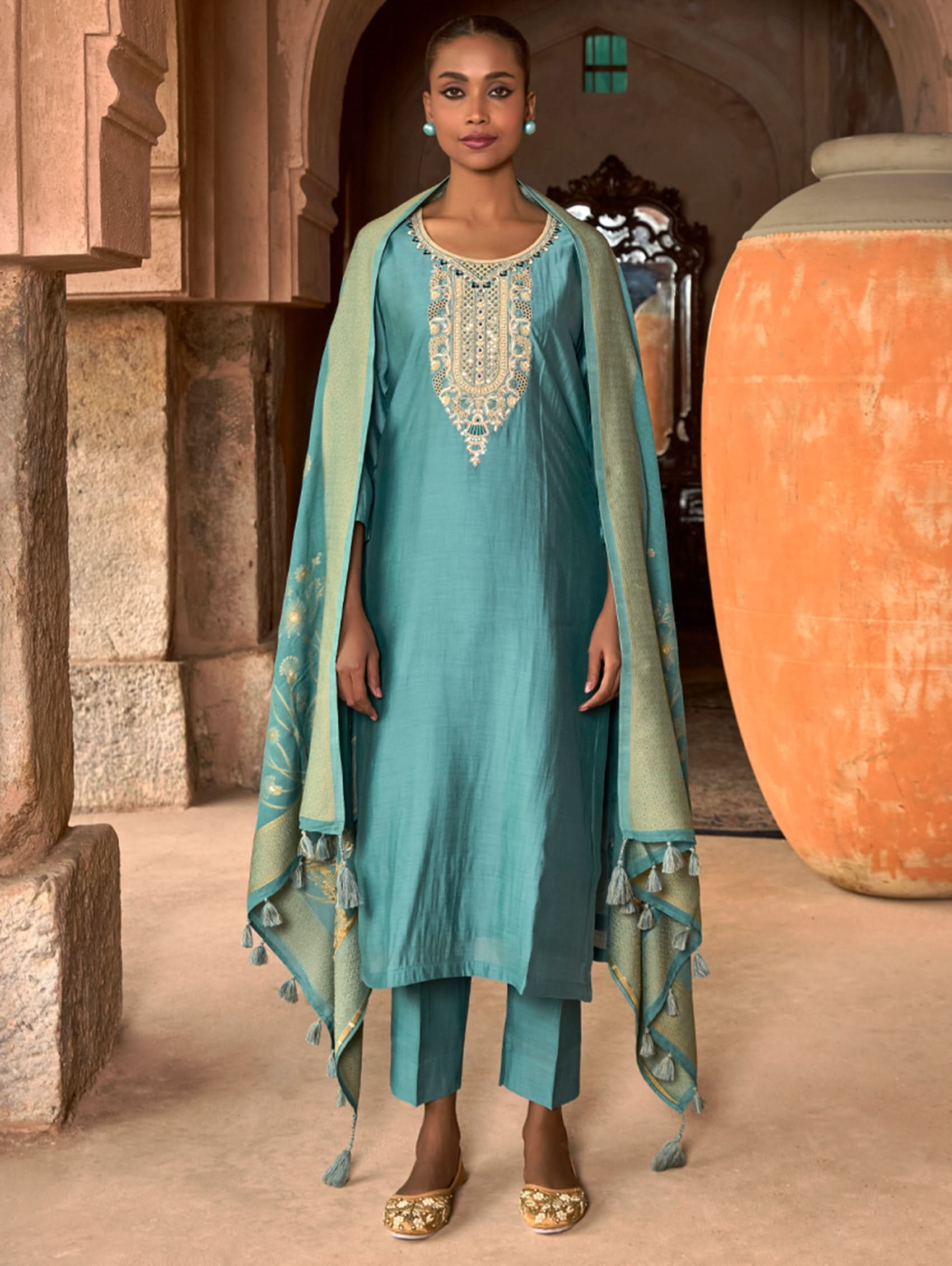 A woman wearing a blue viscose silk suit set with embroidered details, consisting of a kurta, pants, and a dupatta, standing in front of a pot.