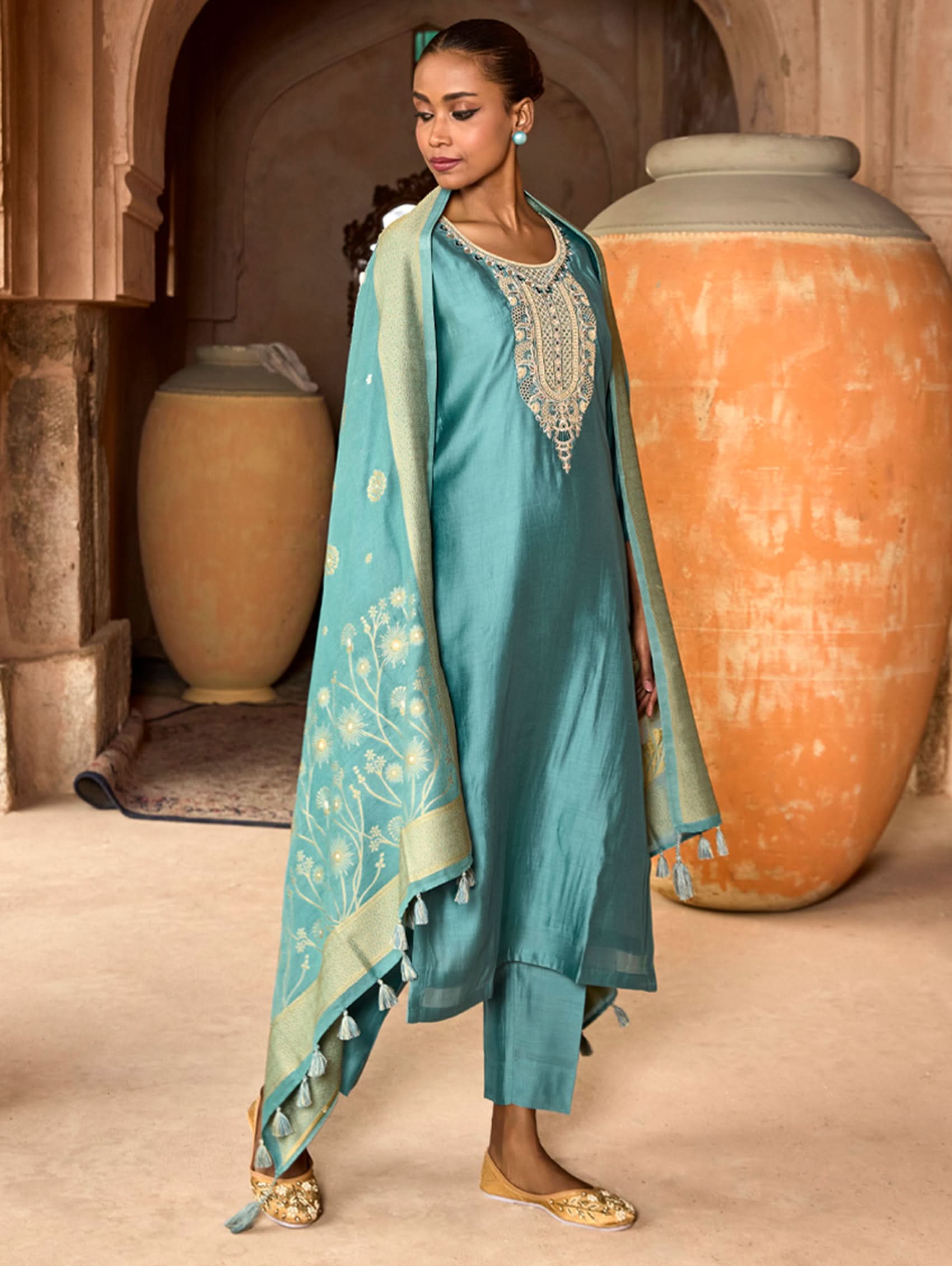 A woman wearing a blue viscose silk suit set with embroidered details, consisting of a kurta, pants, and a dupatta, standing in front of a pot.