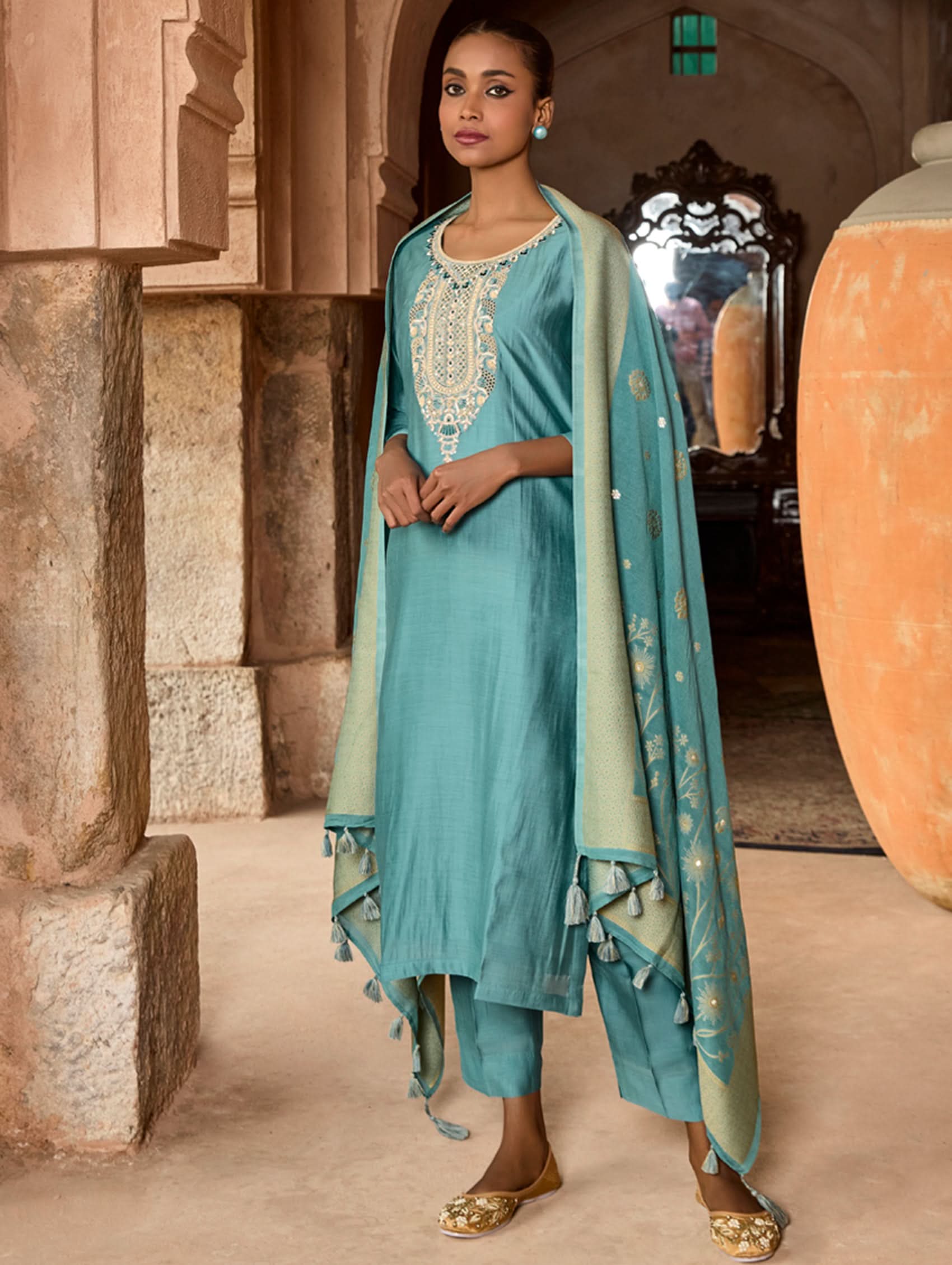 A woman wearing a blue viscose silk suit set with embroidered details, consisting of a kurta, pants, and a dupatta, standing in front of a pot.