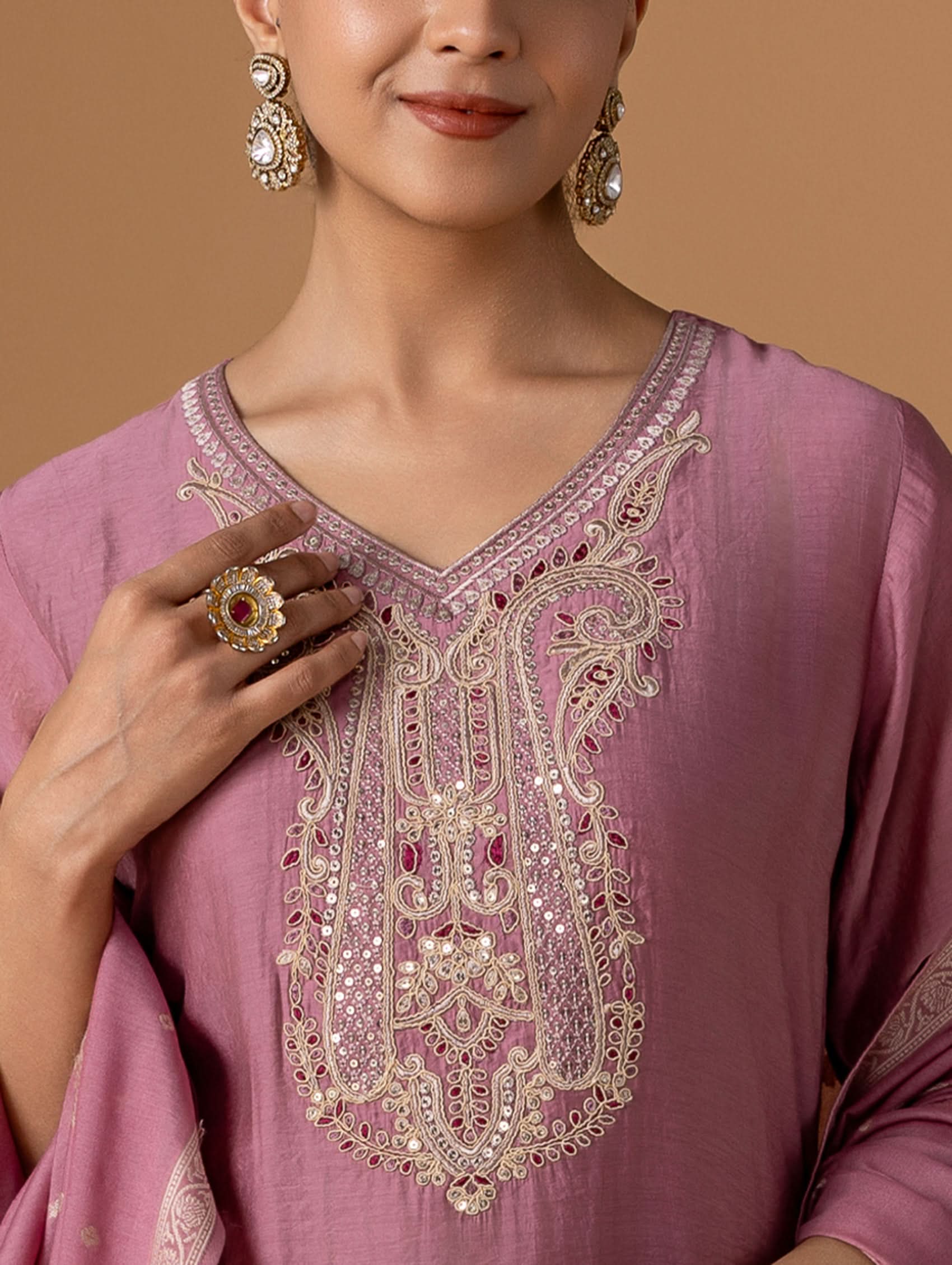 Woman wearing a pink embroidered top with gold jewelry on a brown background