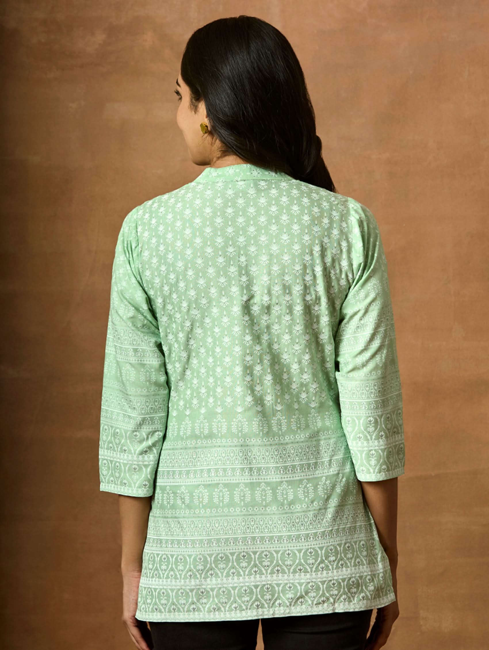 A woman wearing a Green printed viscose rayon tunic with a ban collar and three-fourth sleeves, paired with white trousers.
