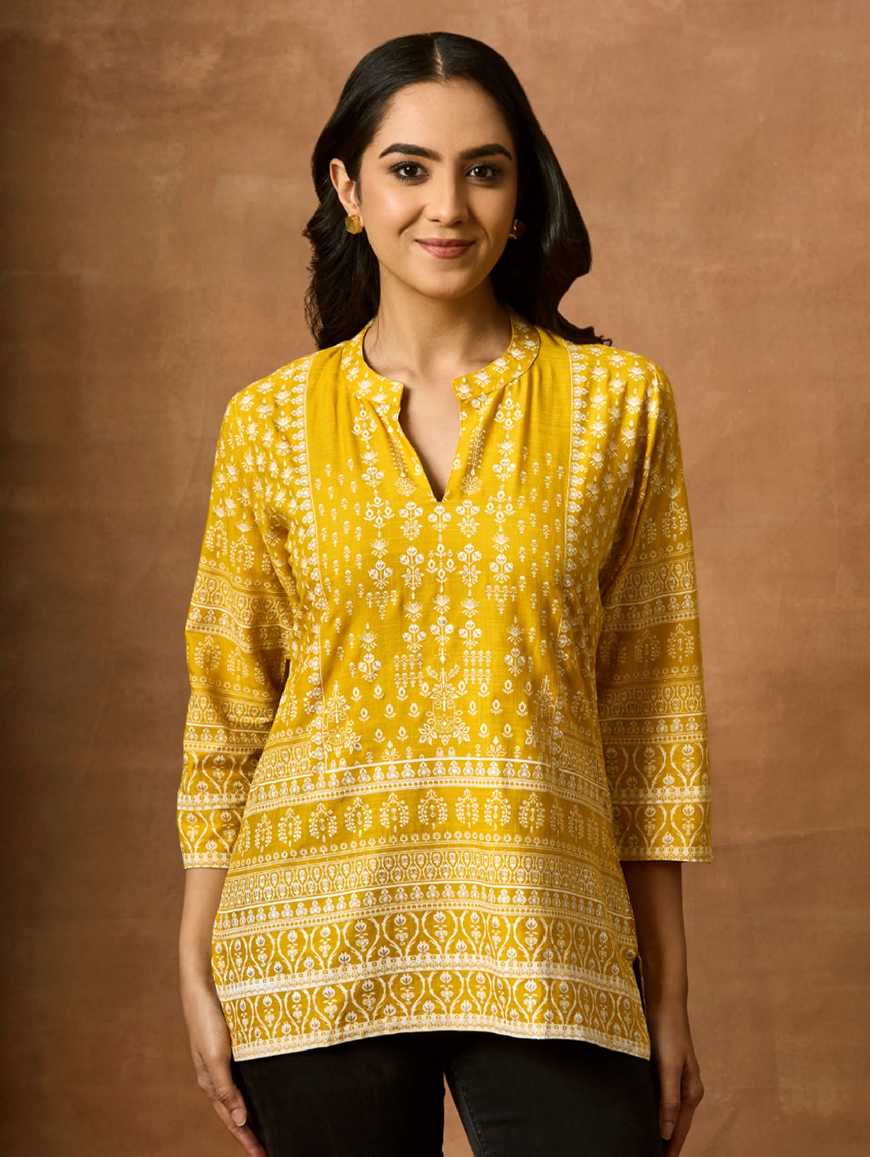 A woman wearing a Yellow printed viscose rayon tunic with a ban collar and three-fourth sleeves, paired with white trousers.