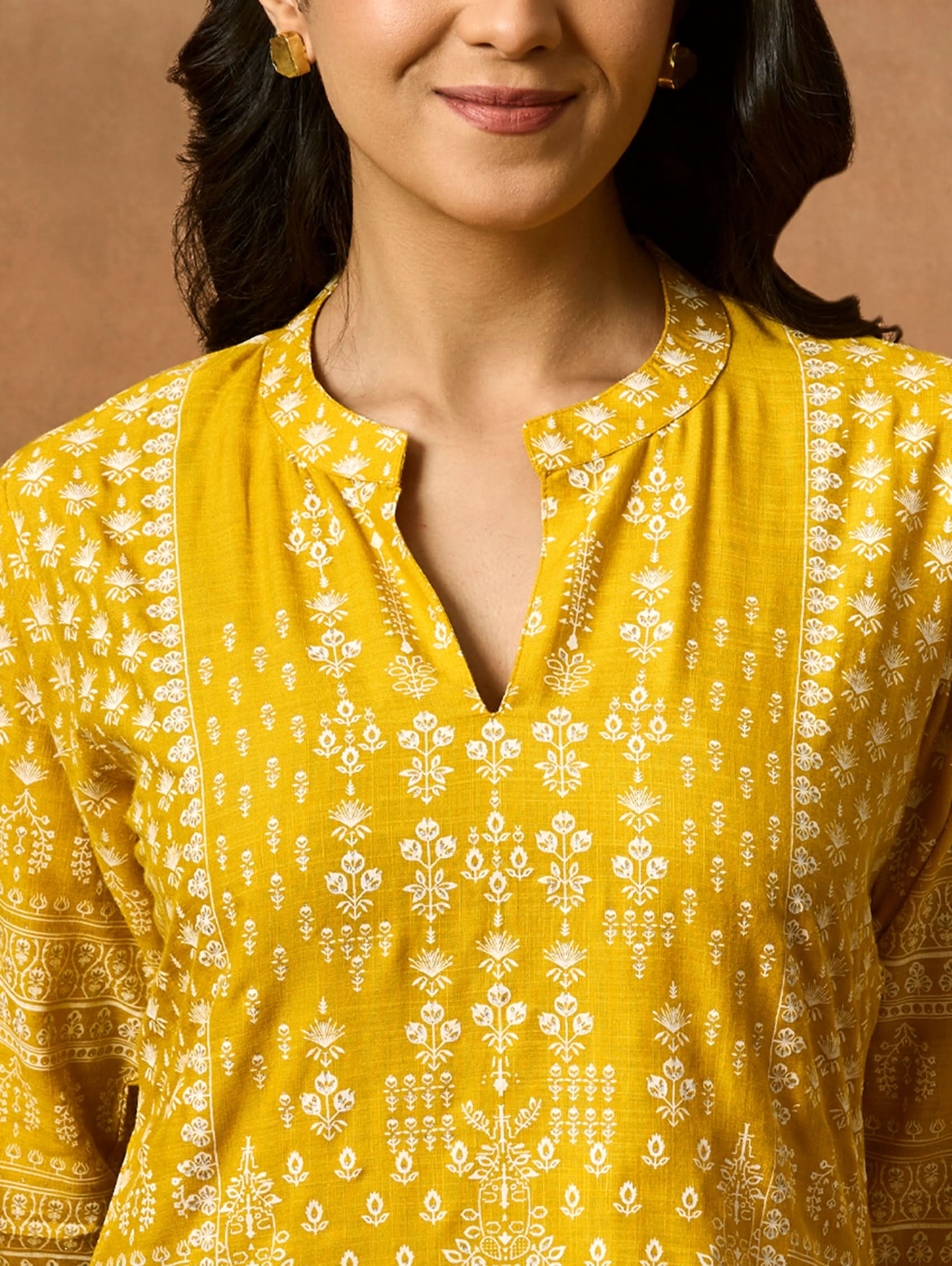 A woman wearing a Yellow printed viscose rayon tunic with a ban collar and three-fourth sleeves, paired with white trousers.