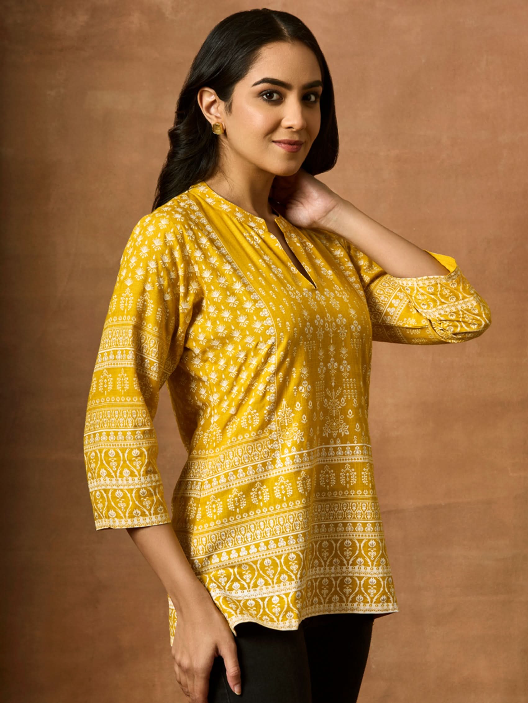 A woman wearing a Yellow printed viscose rayon tunic with a ban collar and three-fourth sleeves, paired with white trousers.