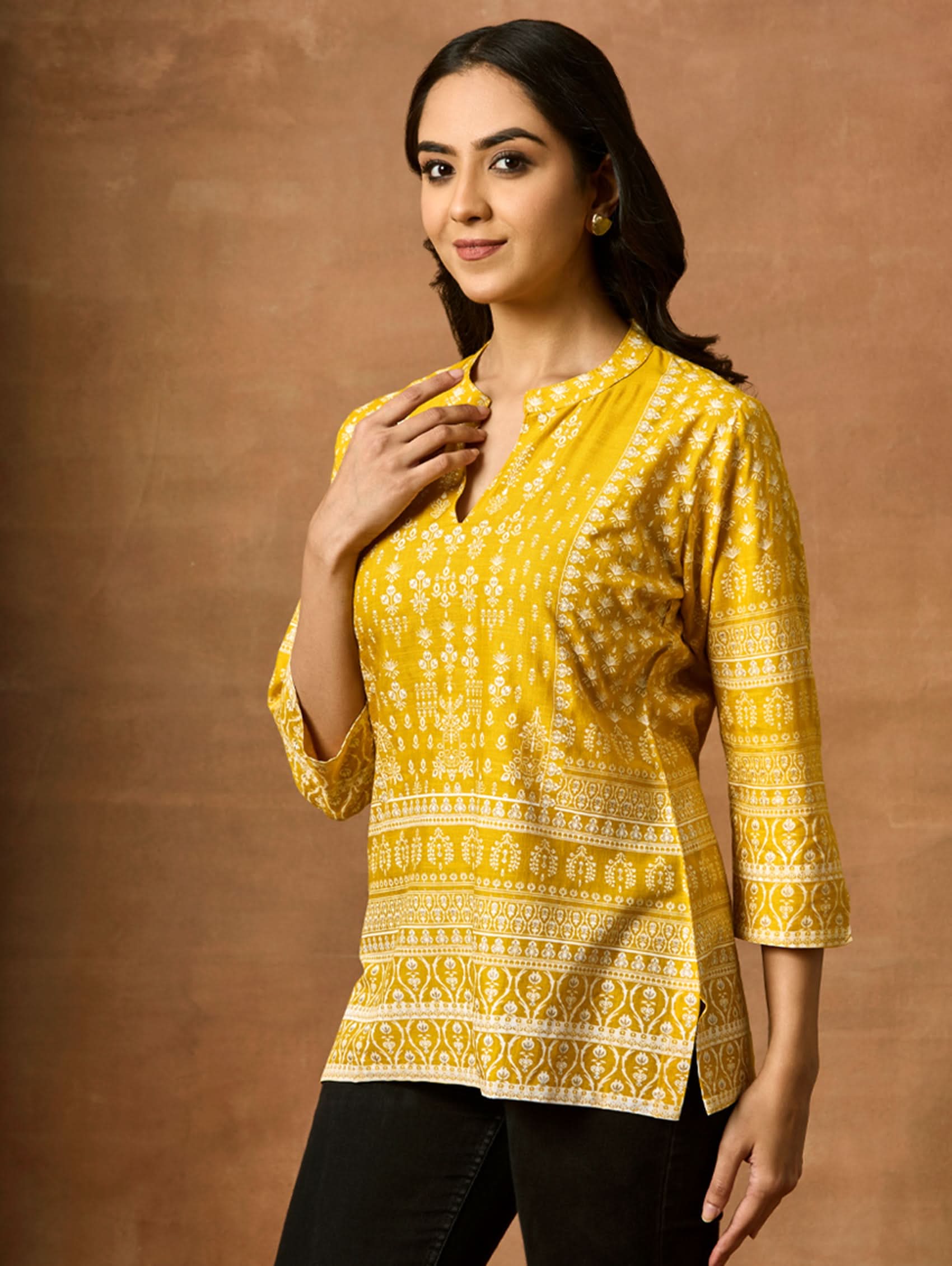 A woman wearing a Yellow printed viscose rayon tunic with a ban collar and three-fourth sleeves, paired with white trousers.