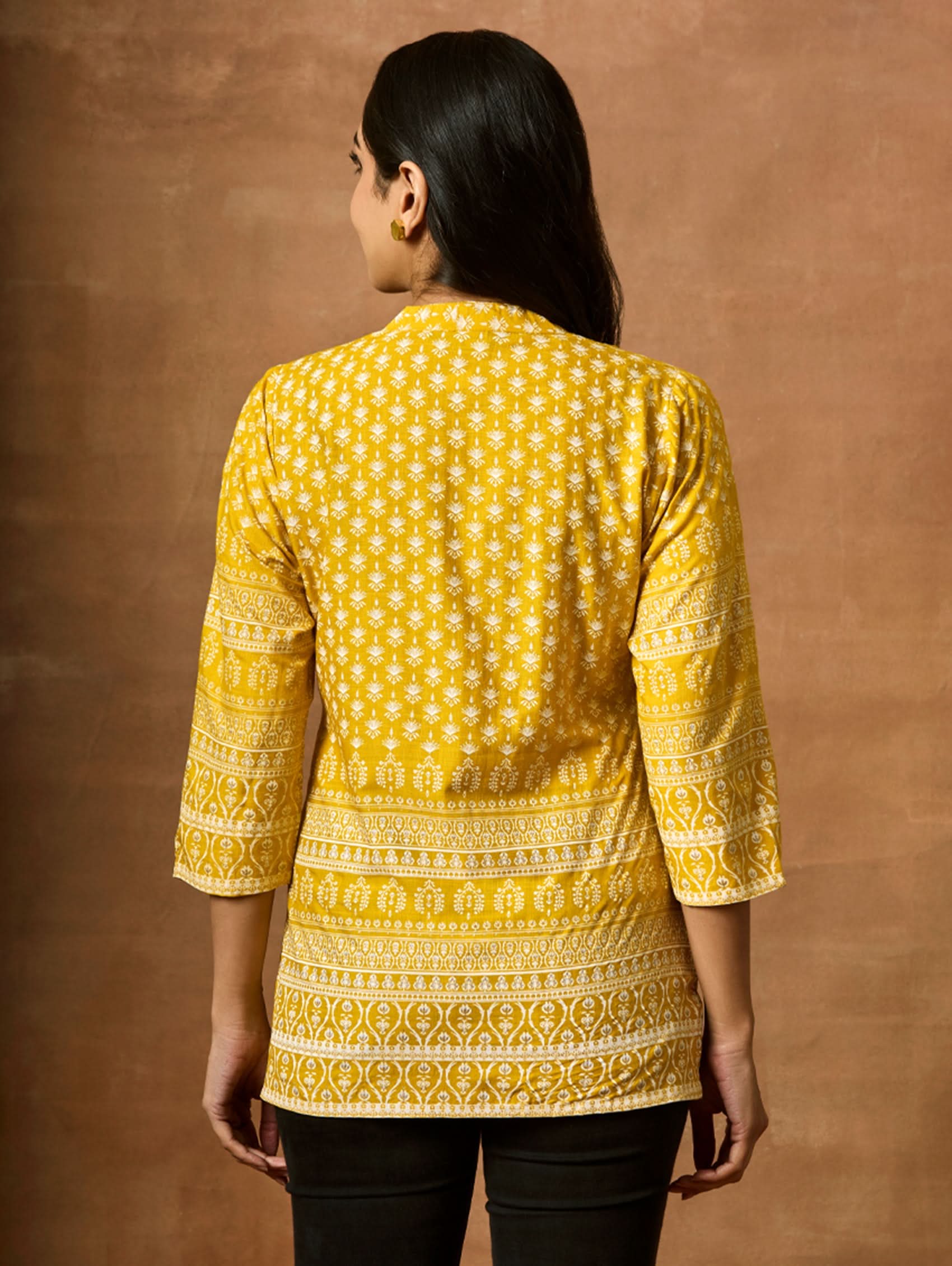 A woman wearing a Yellow printed viscose rayon tunic with a ban collar and three-fourth sleeves, paired with white trousers.
