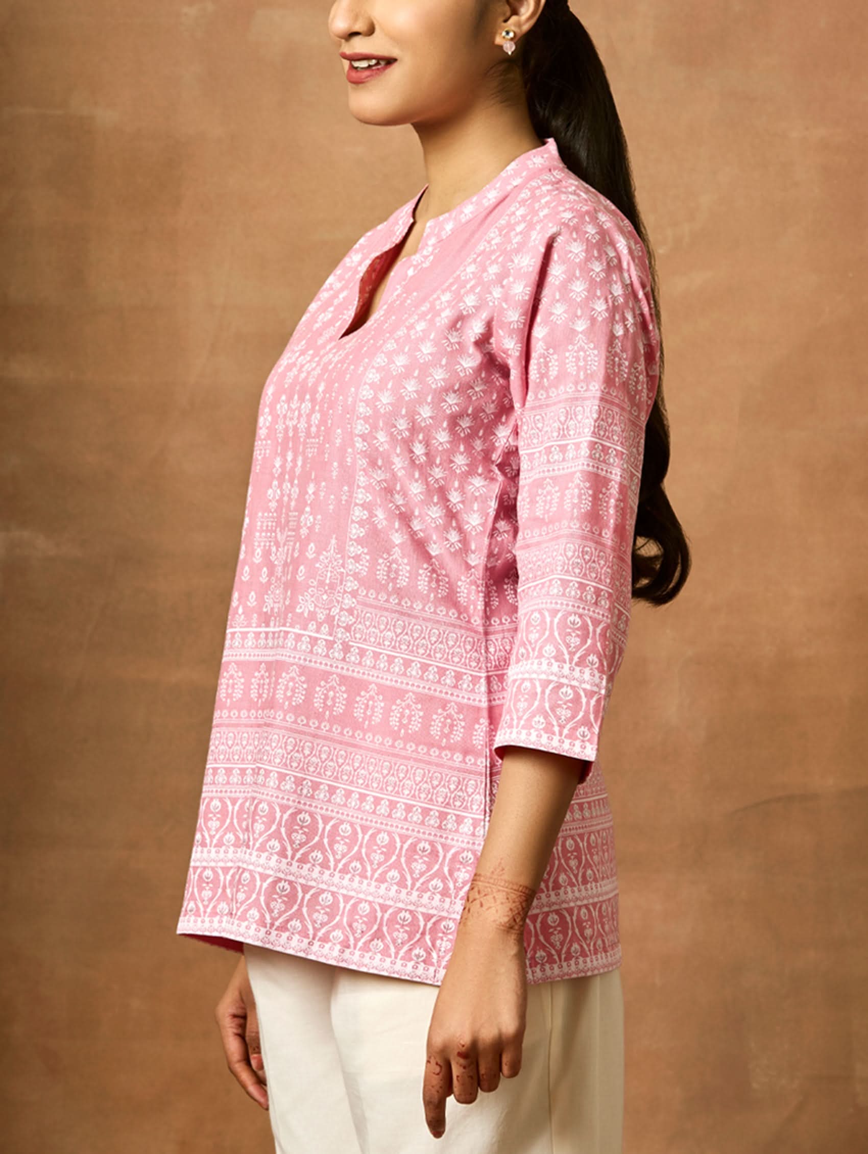 A woman wearing a pink printed viscose rayon tunic with a ban collar and three-fourth sleeves, paired with white trousers.