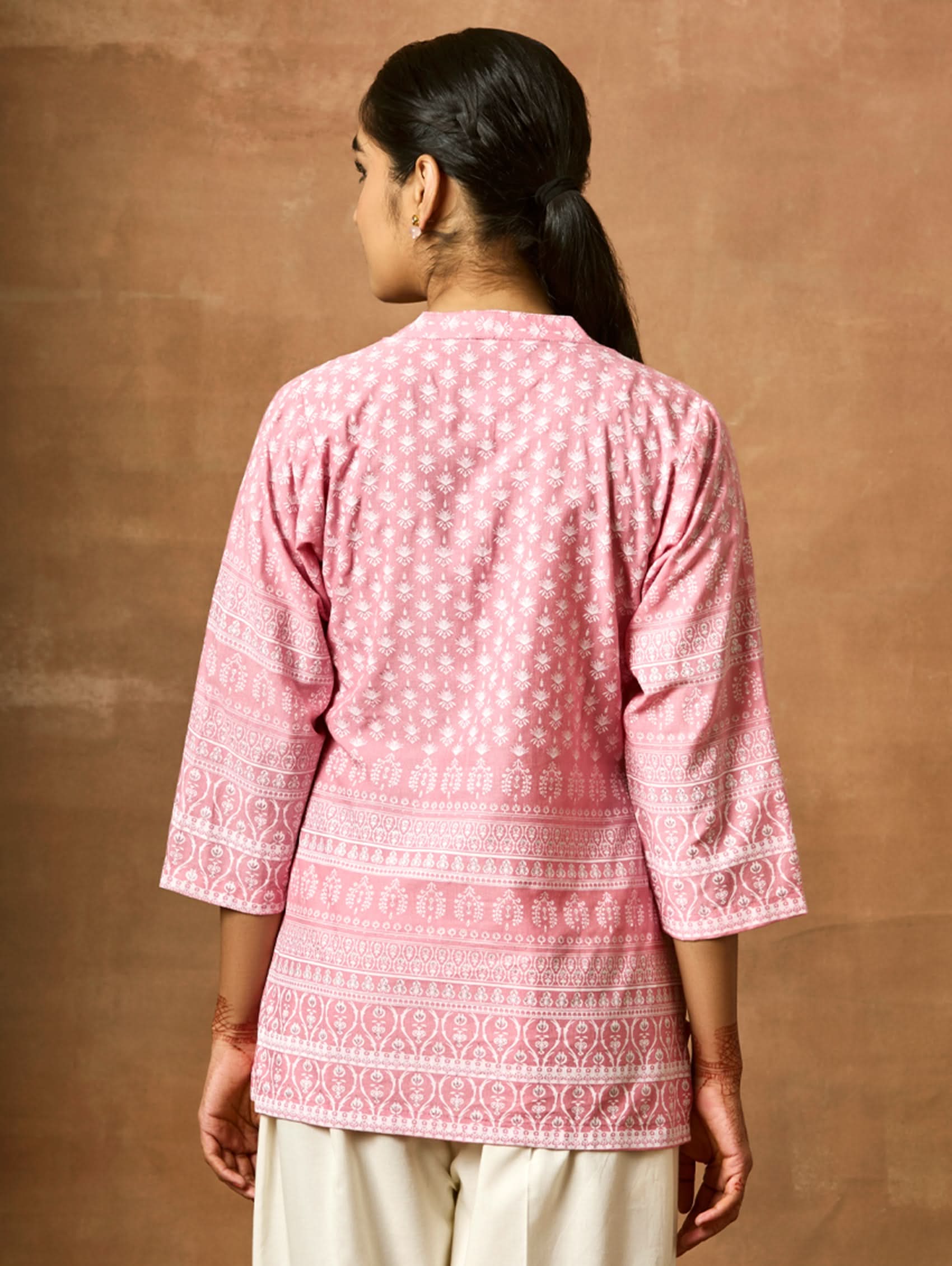 A woman wearing a pink printed viscose rayon tunic with a ban collar and three-fourth sleeves, paired with white trousers.