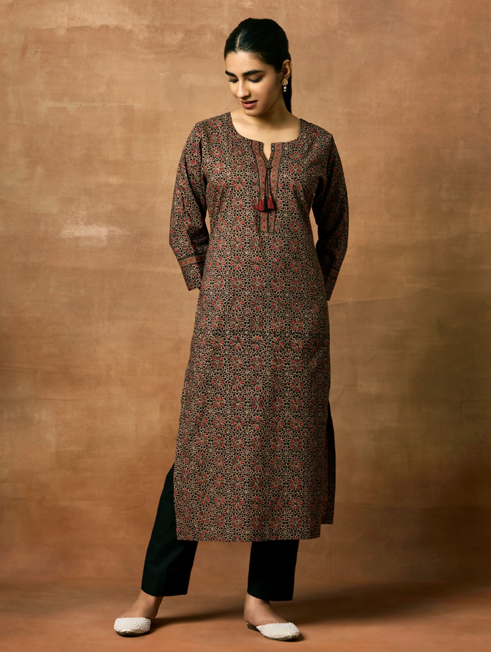 A woman plus-size model wearing a knee-length cotton kurta with a round neckline, three-fourth sleeves, and side slits. The kurta has a round neckline with tassels and is printed with a small floral pattern.