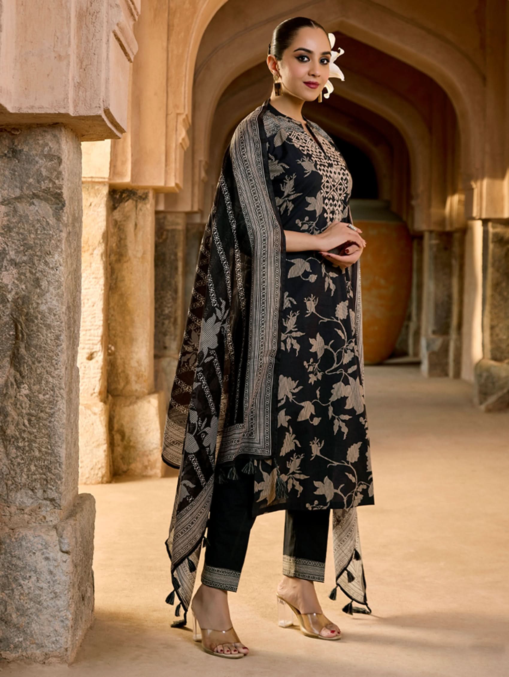 A woman wearing a floral cotton suit set with embroidered details,and printed consisting of a kurta with band collar, pants, and a printed weave silk dupatta.