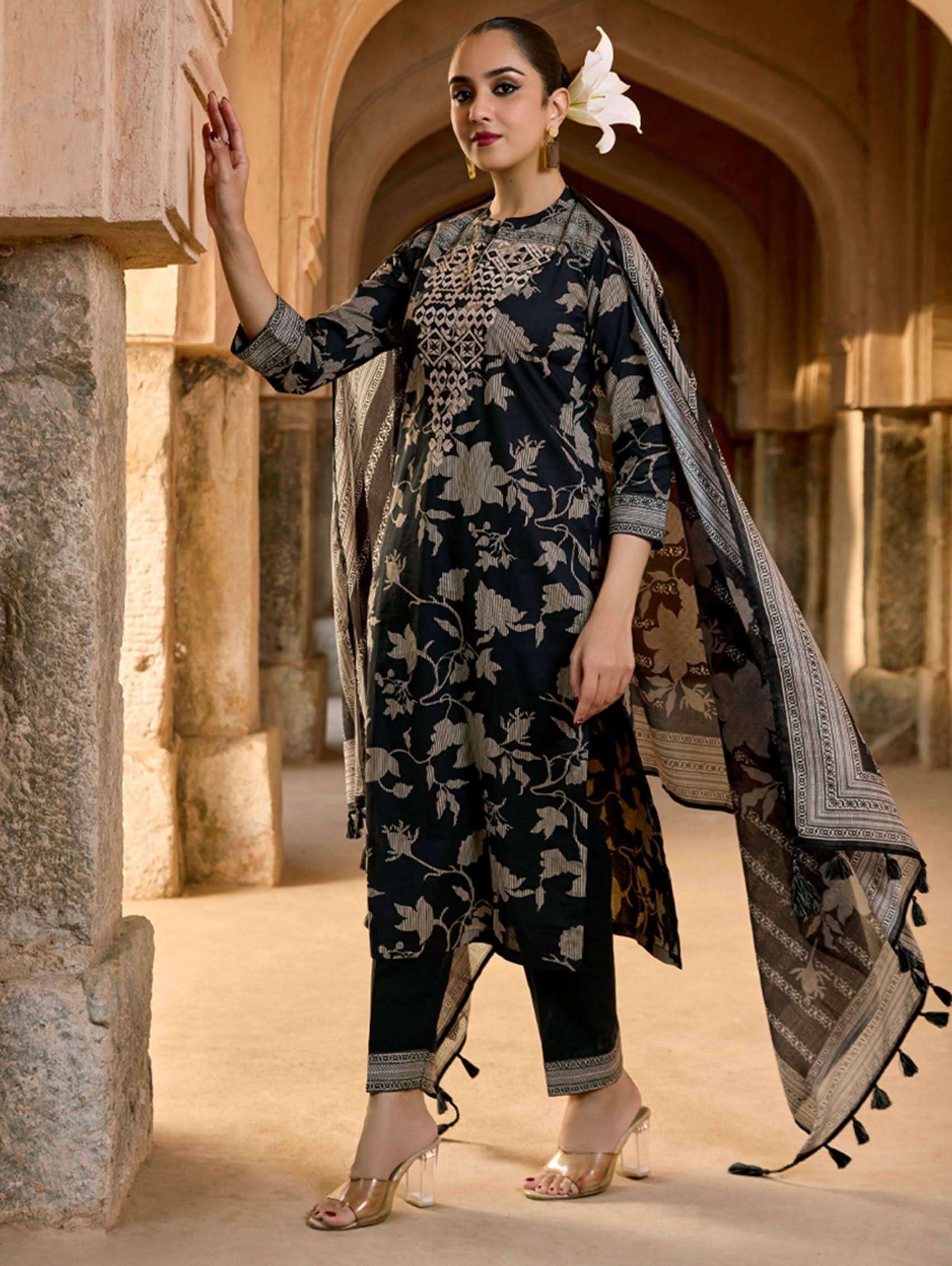 A woman wearing a floral cotton suit set with embroidered details,and printed consisting of a kurta with band collar, pants, and a printed weave silk dupatta.