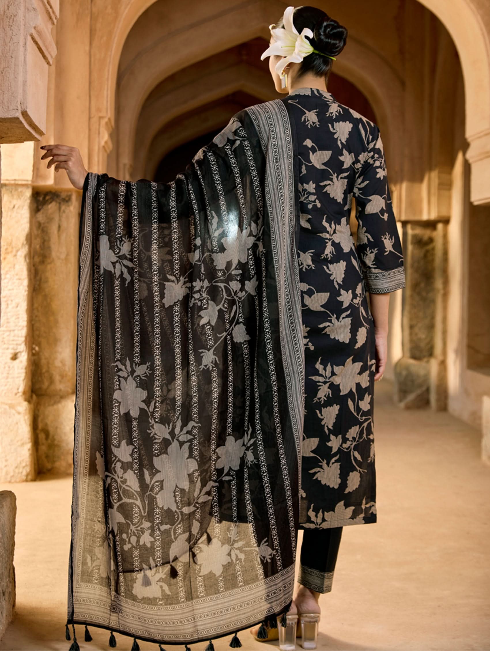 A woman wearing a floral cotton suit set with embroidered details,and printed consisting of a kurta with band collar, pants, and a printed weave silk dupatta.