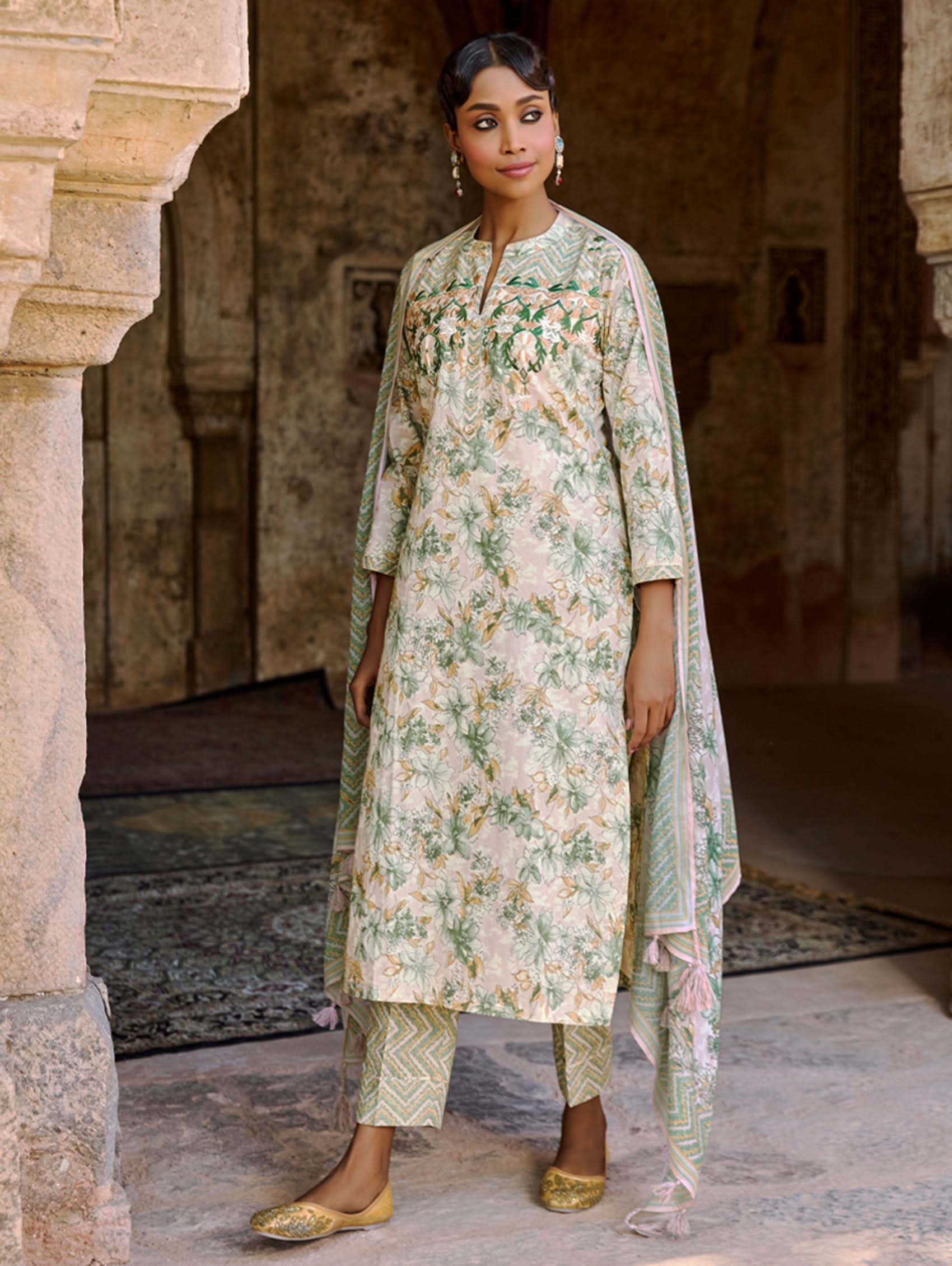 A woman wearing a floral cotton suit set with embroidered details,and printed consisting of a kurta with band collor, pants, and a printed weave silk dupatta.
