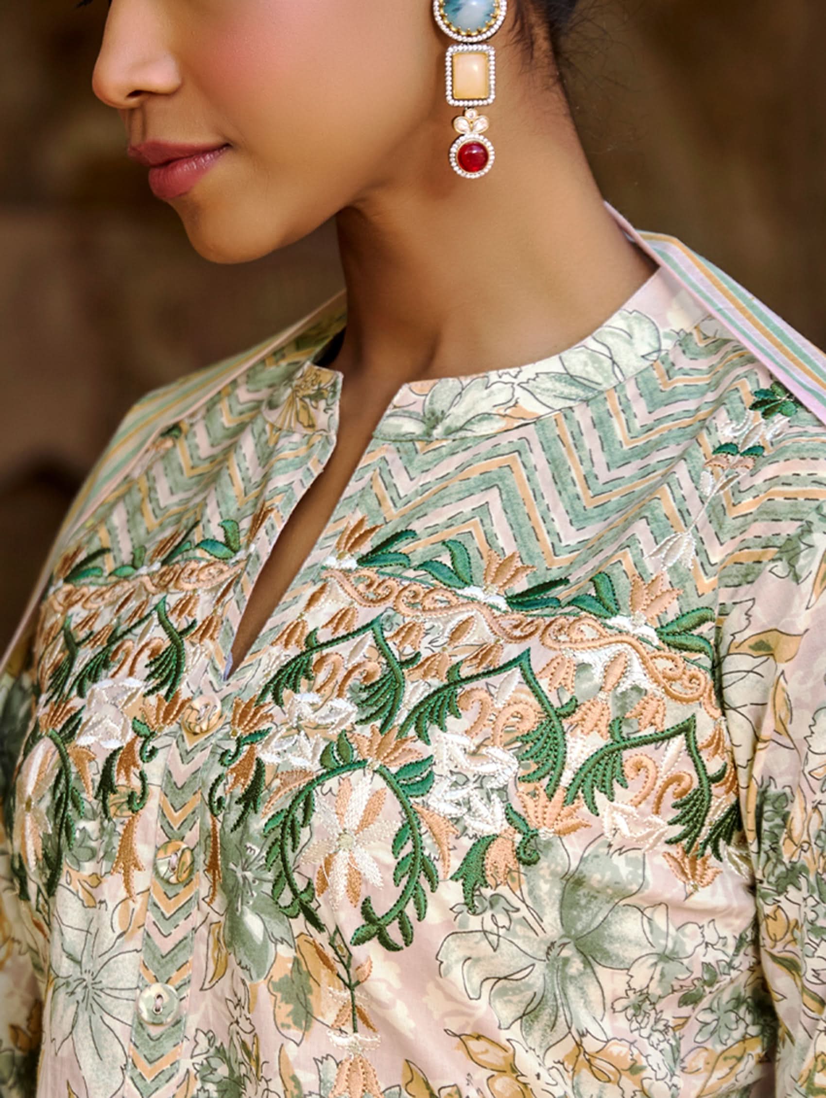 A woman wearing a floral cotton suit set with embroidered details,and printed consisting of a kurta with band collor, pants, and a printed weave silk dupatta.