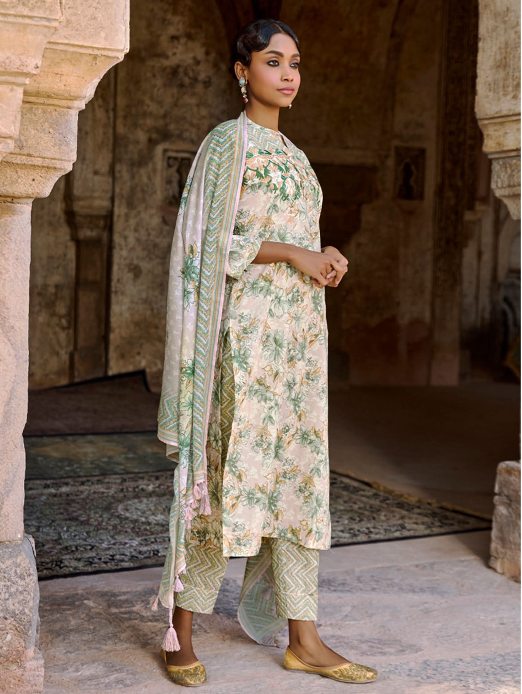 A woman wearing a floral cotton suit set with embroidered details,and printed consisting of a kurta with band collor, pants, and a printed weave silk dupatta.