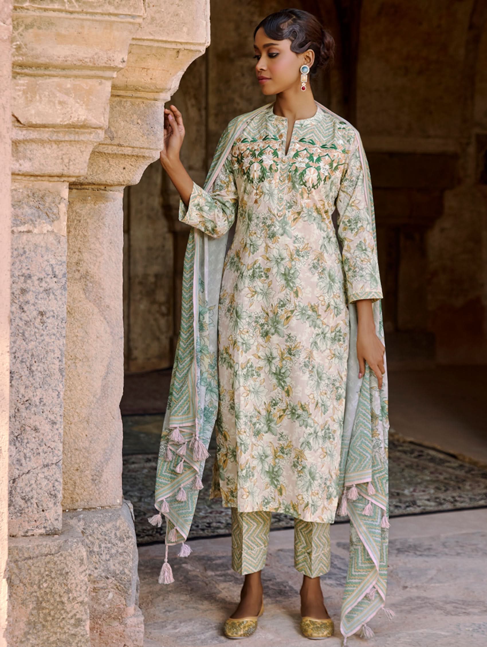 A woman wearing a floral cotton suit set with embroidered details,and printed consisting of a kurta with band collor, pants, and a printed weave silk dupatta.