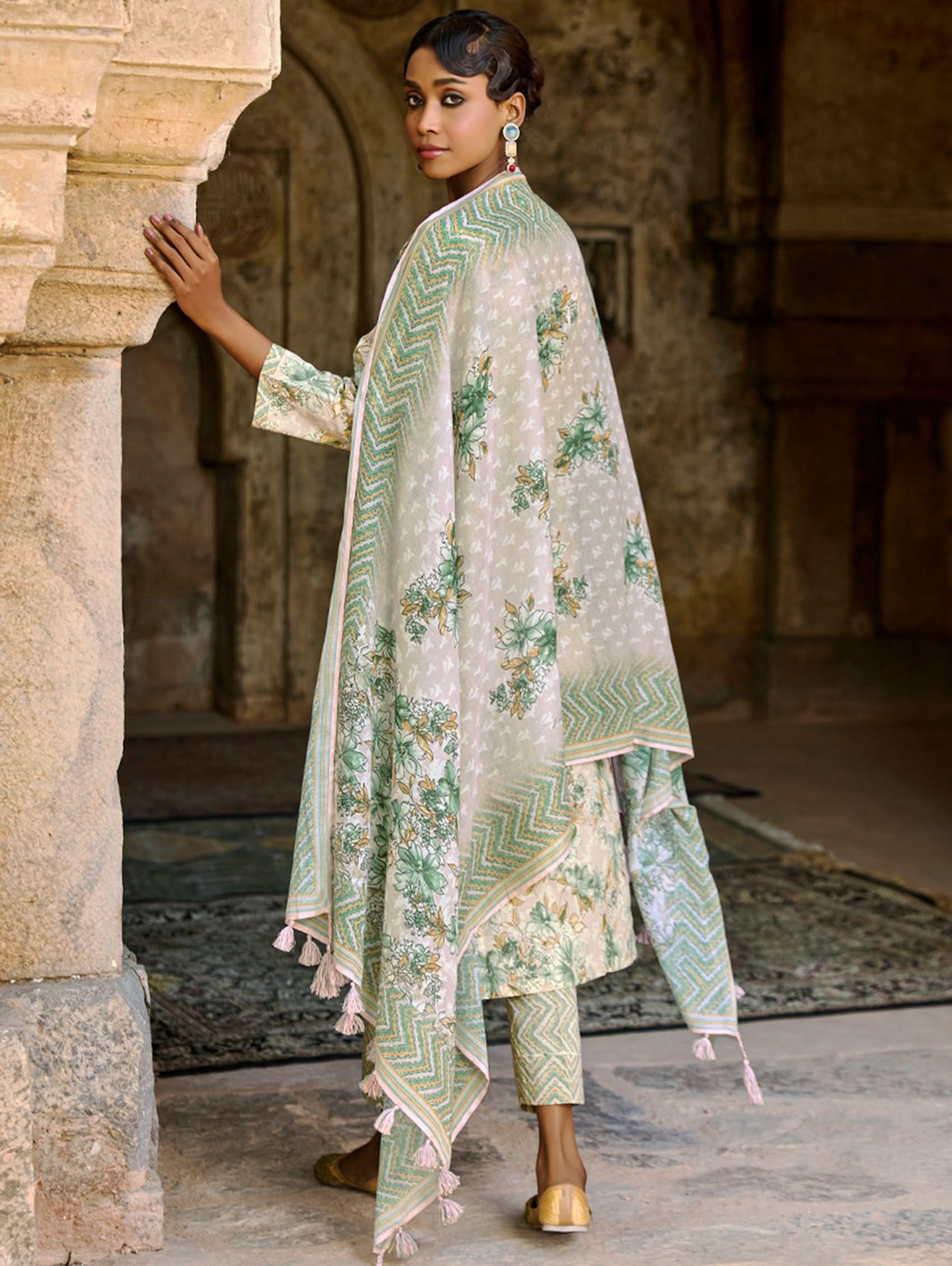 A woman wearing a floral cotton suit set with embroidered details,and printed consisting of a kurta with band collor, pants, and a printed weave silk dupatta.