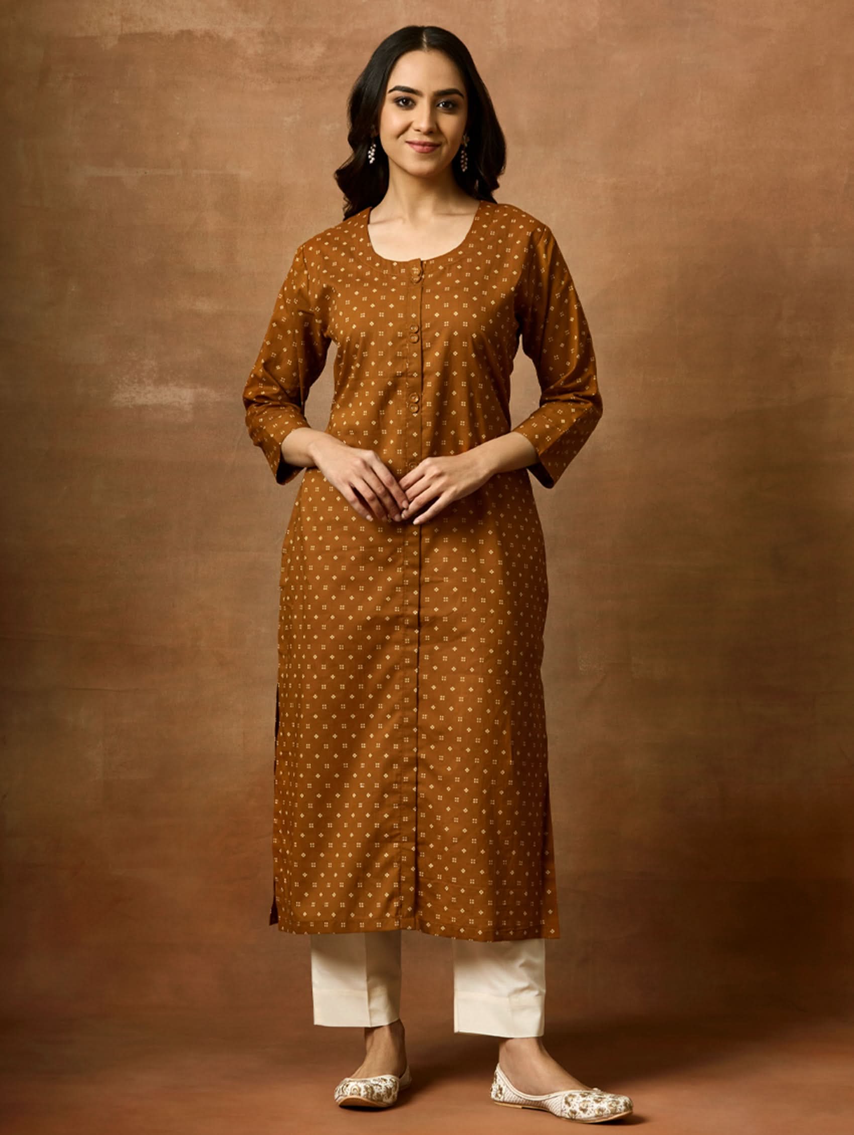 A woman standing against a brown background, wearing a brown printed pure cotton kurta with a round neckline, three-fourth sleeves, and side slits. She is also wearing light-colored pants and white sandals.