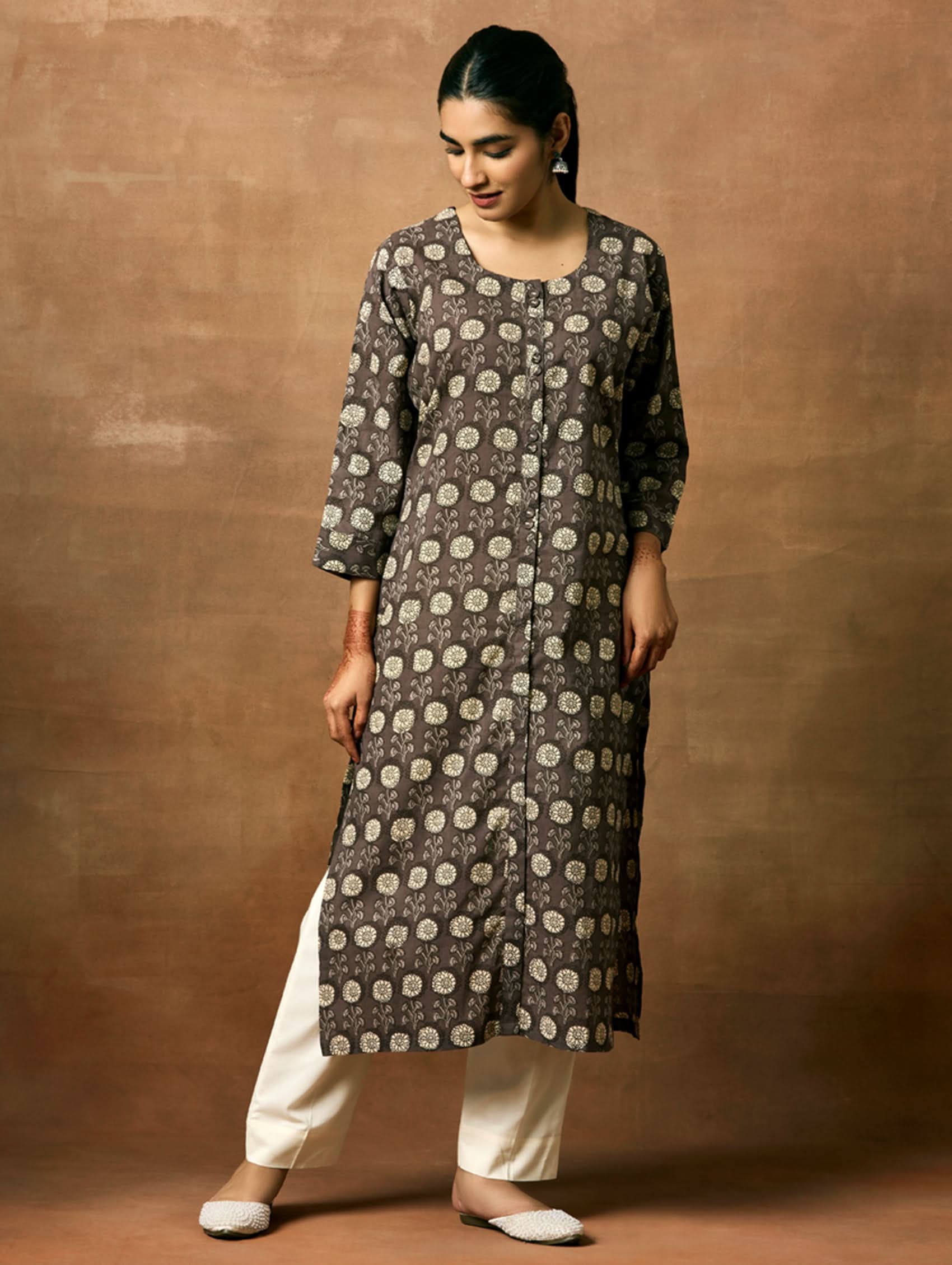 A woman plus size model wearing a printed pure cotton kurta with round neckline, three-fourth sleeves, and side slits. The kurta is in shades of gray and the model is paired with light pants and white shoes.