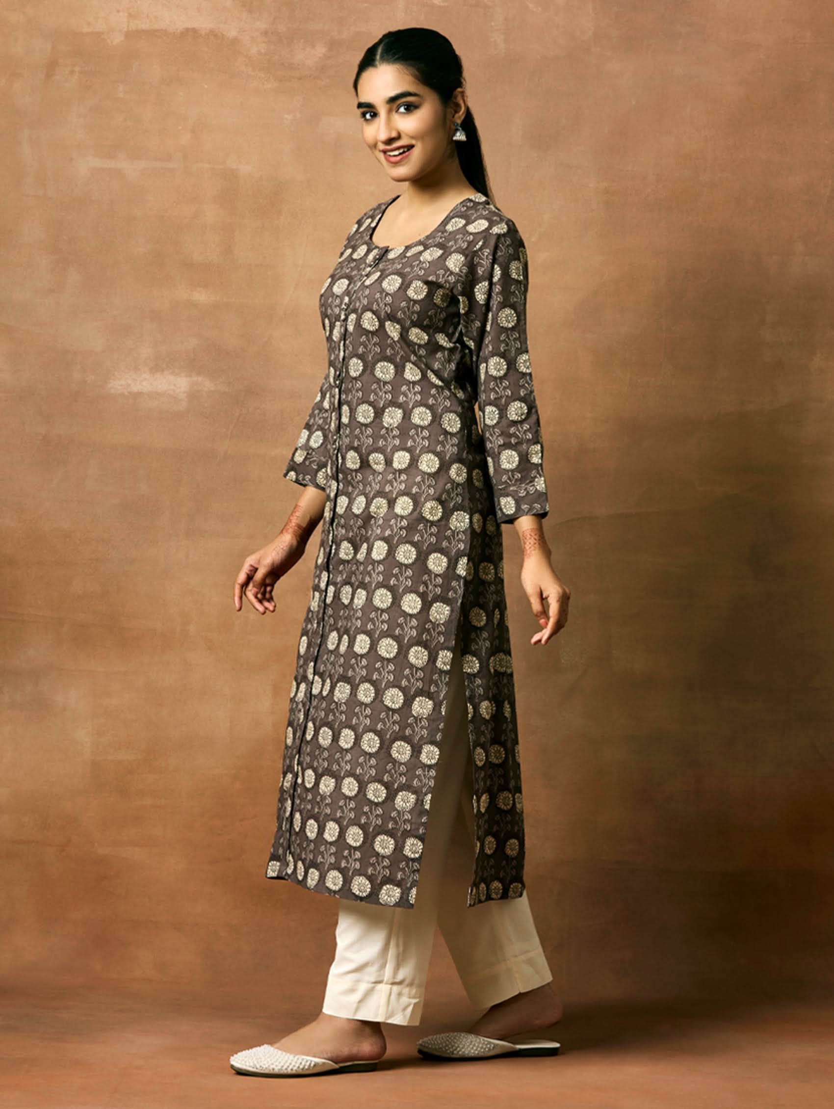 A woman plus size model wearing a printed pure cotton kurta with round neckline, three-fourth sleeves, and side slits. The kurta is in shades of gray and the model is paired with light pants and white shoes.