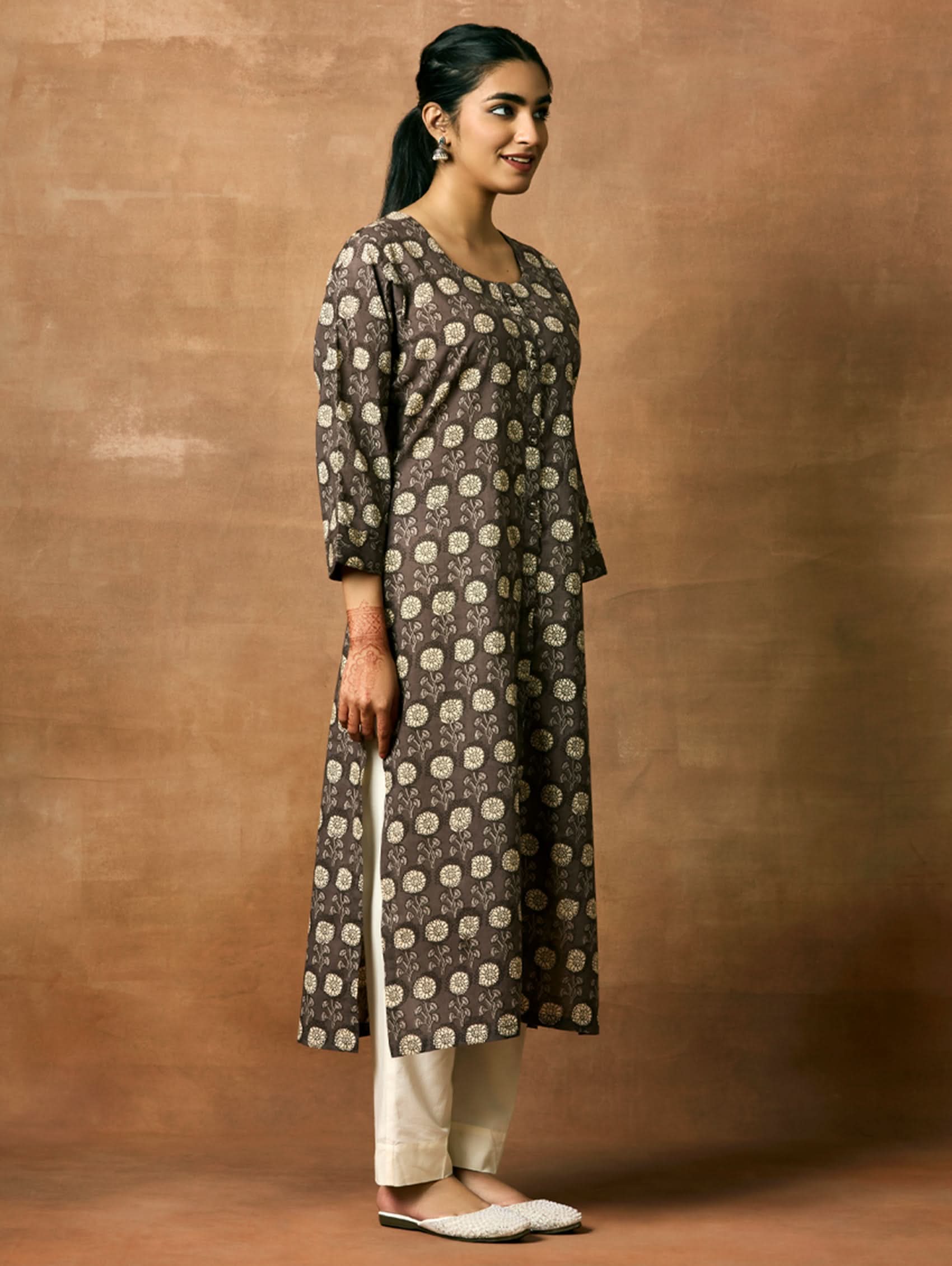 A woman plus size model wearing a printed pure cotton kurta with round neckline, three-fourth sleeves, and side slits. The kurta is in shades of gray and the model is paired with light pants and white shoes.