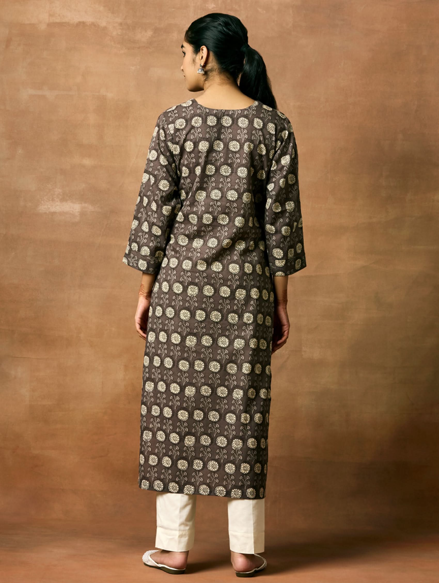 A woman plus size model wearing a printed pure cotton kurta with round neckline, three-fourth sleeves, and side slits. The kurta is in shades of gray and the model is paired with light pants and white shoes.