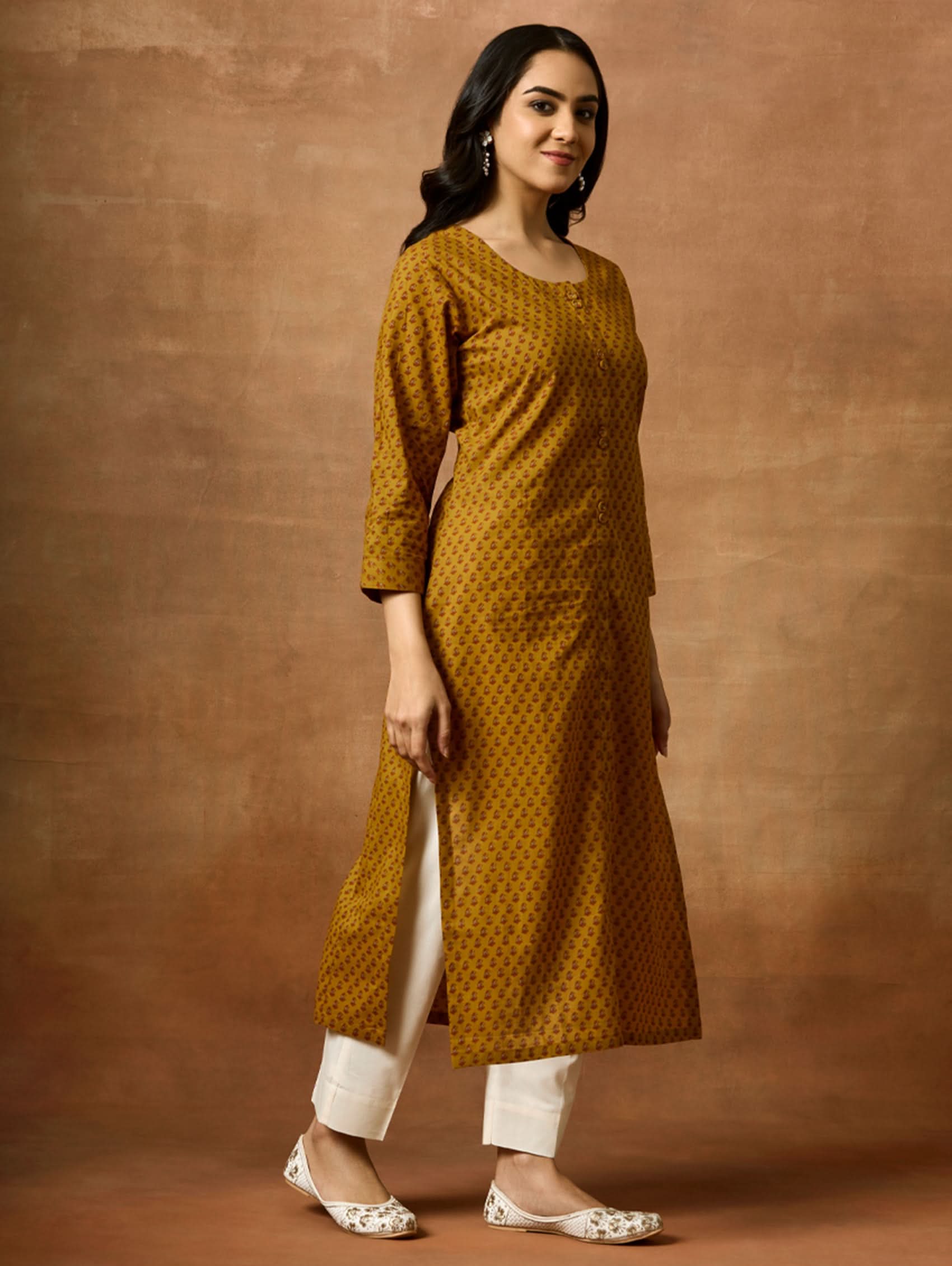 Woman wearing a mustard yellow patterned kurta and white pants against a brown background.