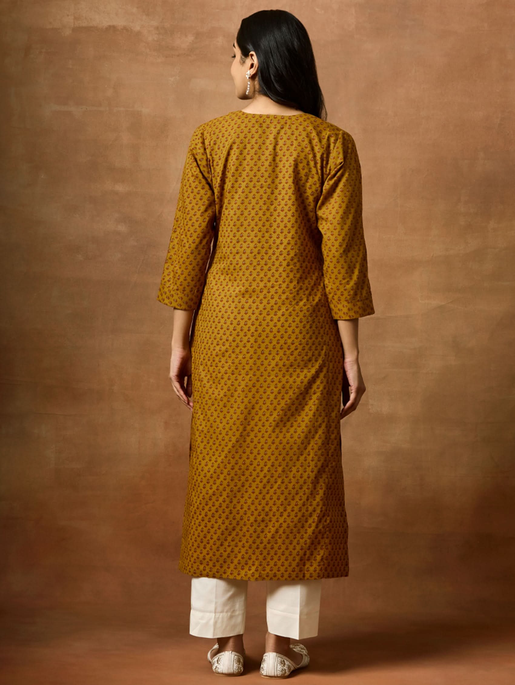 Woman wearing a mustard yellow patterned kurta and white pants against a brown background.