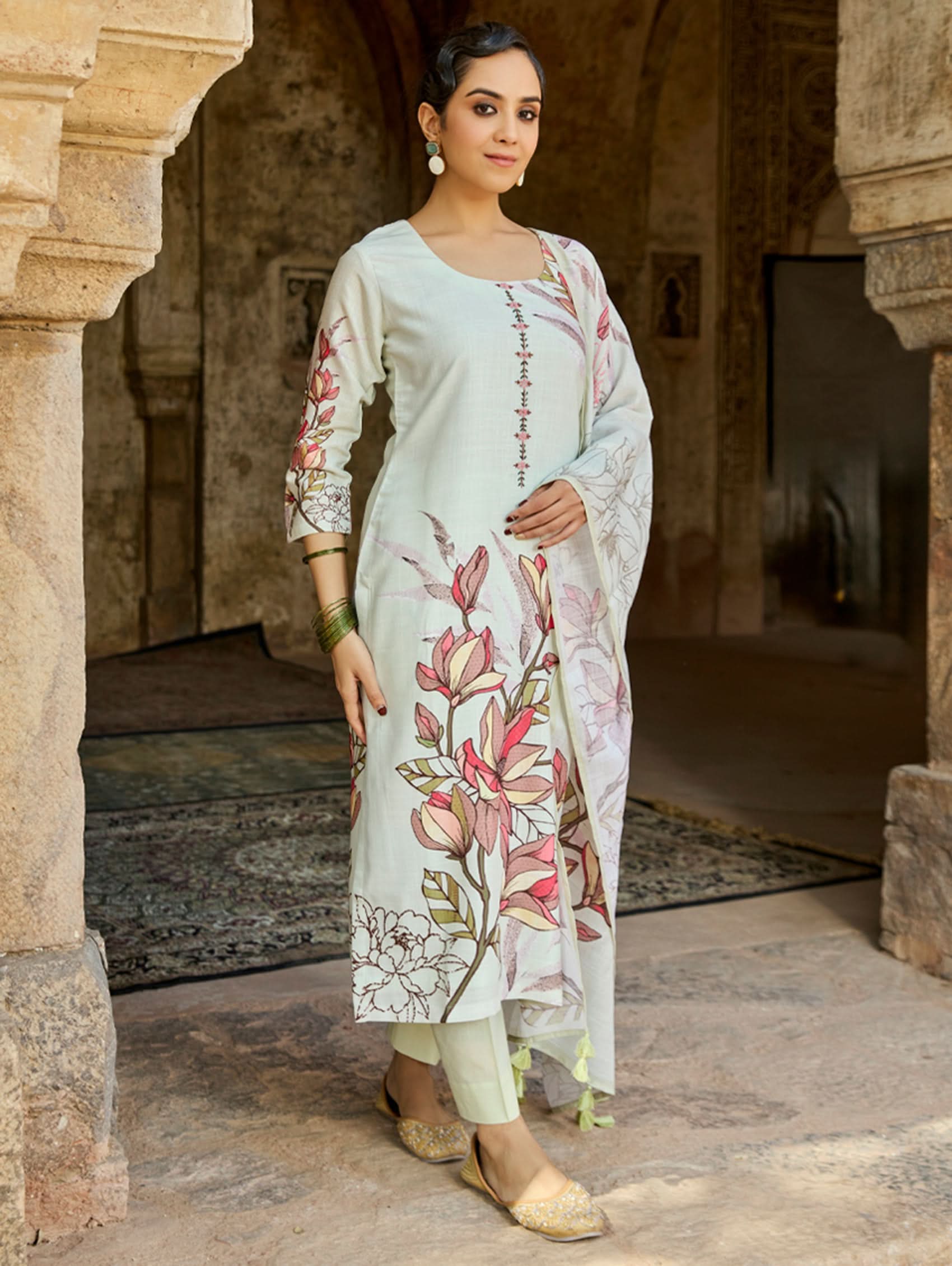 A woman standing in an archway, wearing a digitally printed cotton linen suit set with a round neckline kurta, three-fourth sleeves, side slits, and a dupatta with a floral pattern.