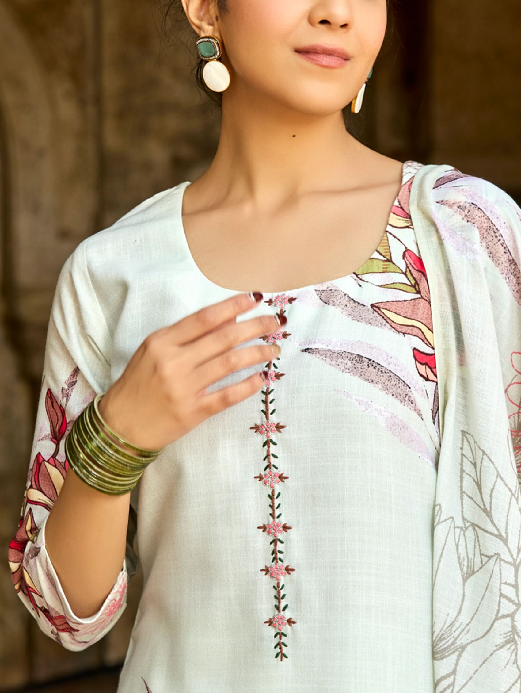 A woman standing in an archway, wearing a digitally printed cotton linen suit set with a round neckline kurta, three-fourth sleeves, side slits, and a dupatta with a floral pattern.