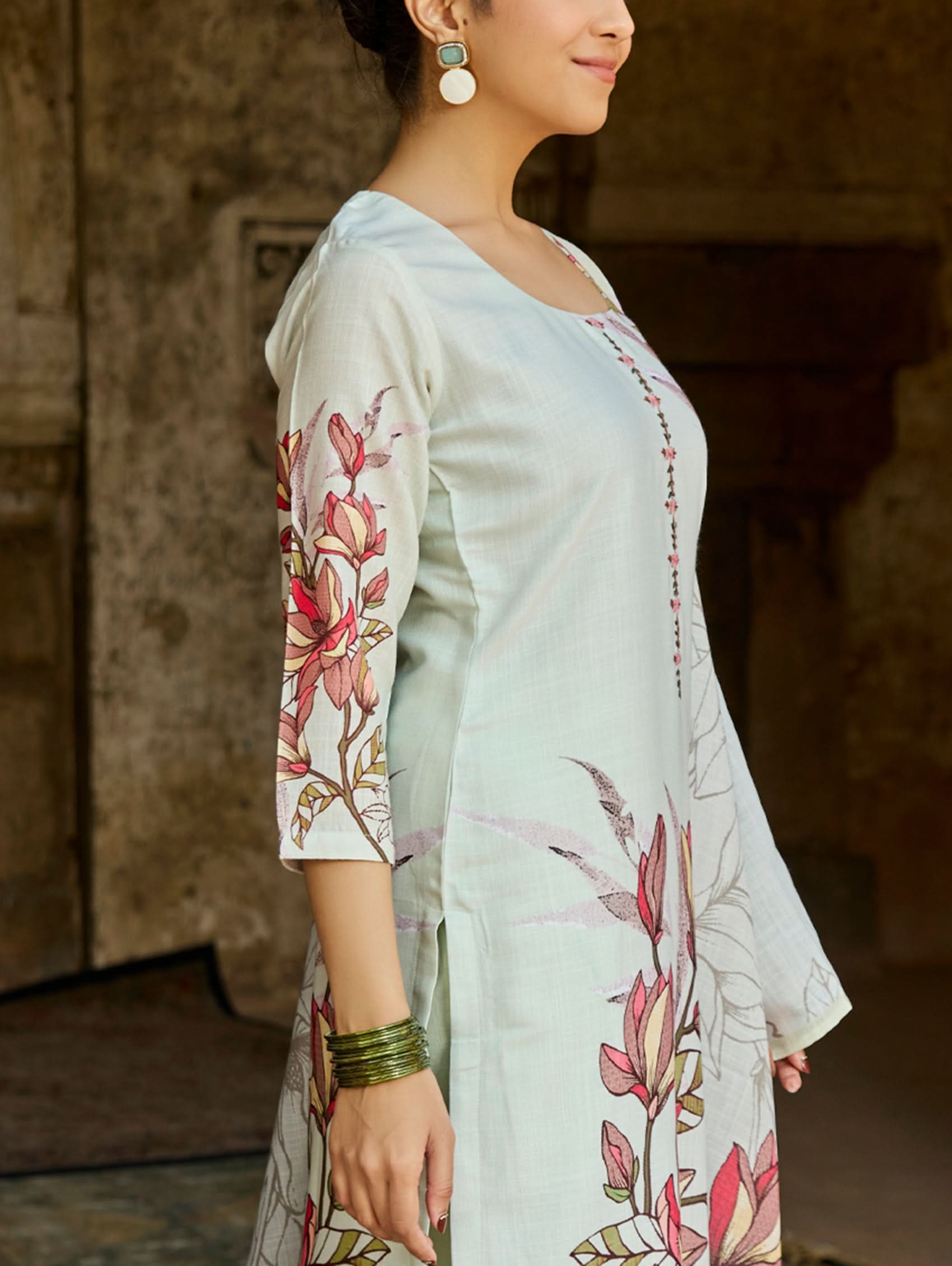 A woman standing in an archway, wearing a digitally printed cotton linen suit set with a round neckline kurta, three-fourth sleeves, side slits, and a dupatta with a floral pattern.