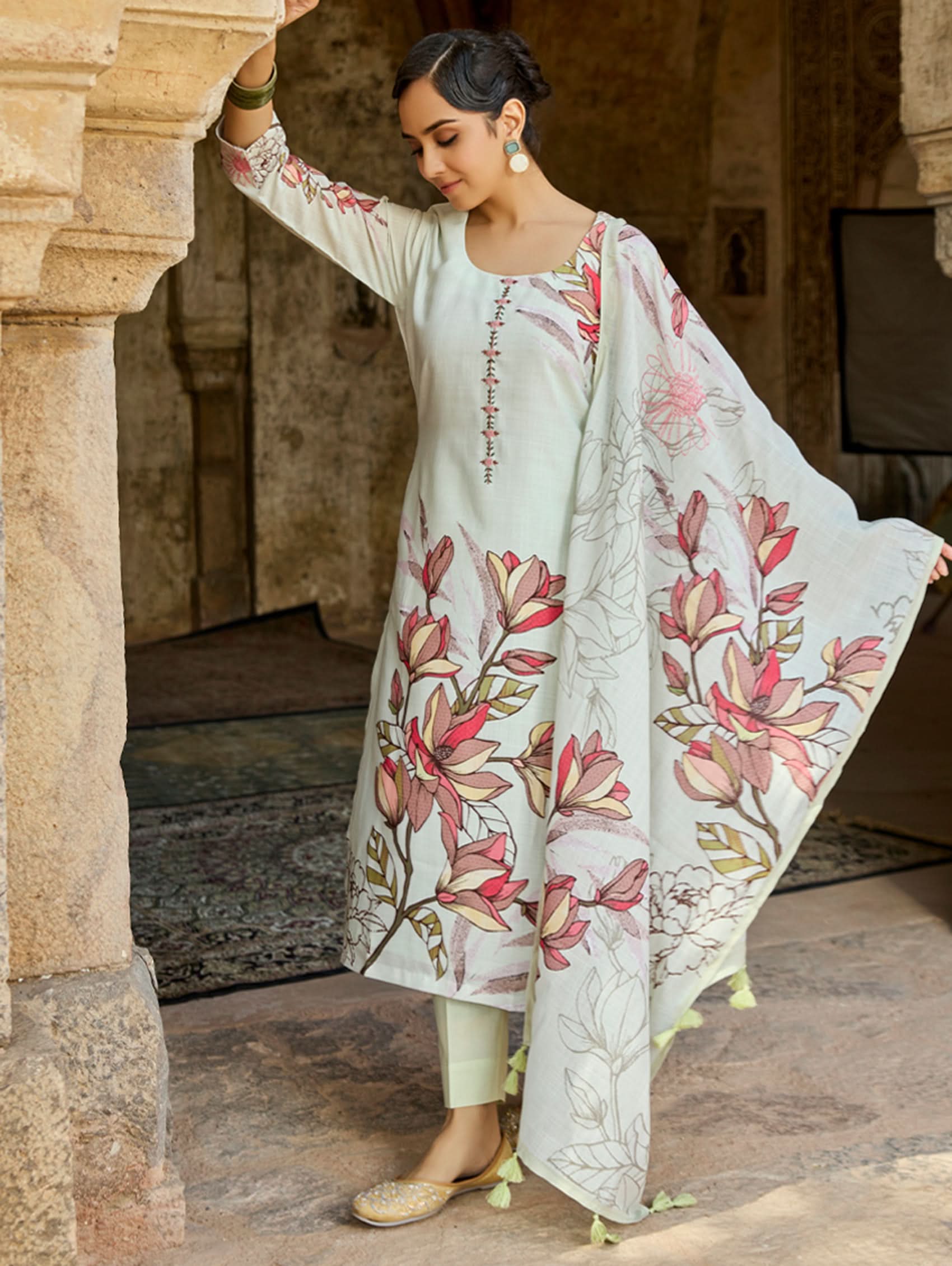 A woman standing in an archway, wearing a digitally printed cotton linen suit set with a round neckline kurta, three-fourth sleeves, side slits, and a dupatta with a floral pattern.