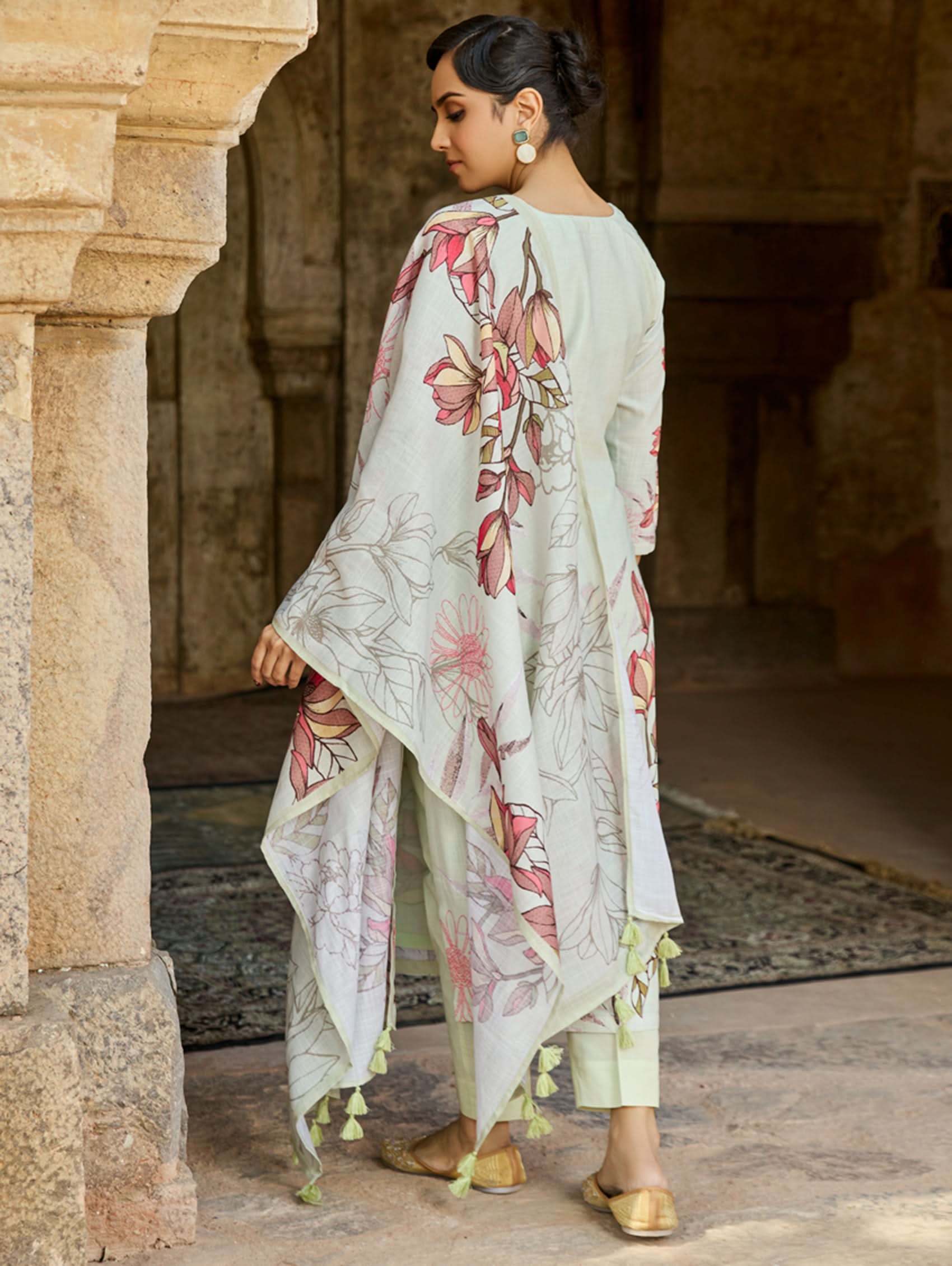 A woman standing in an archway, wearing a digitally printed cotton linen suit set with a round neckline kurta, three-fourth sleeves, side slits, and a dupatta with a floral pattern.