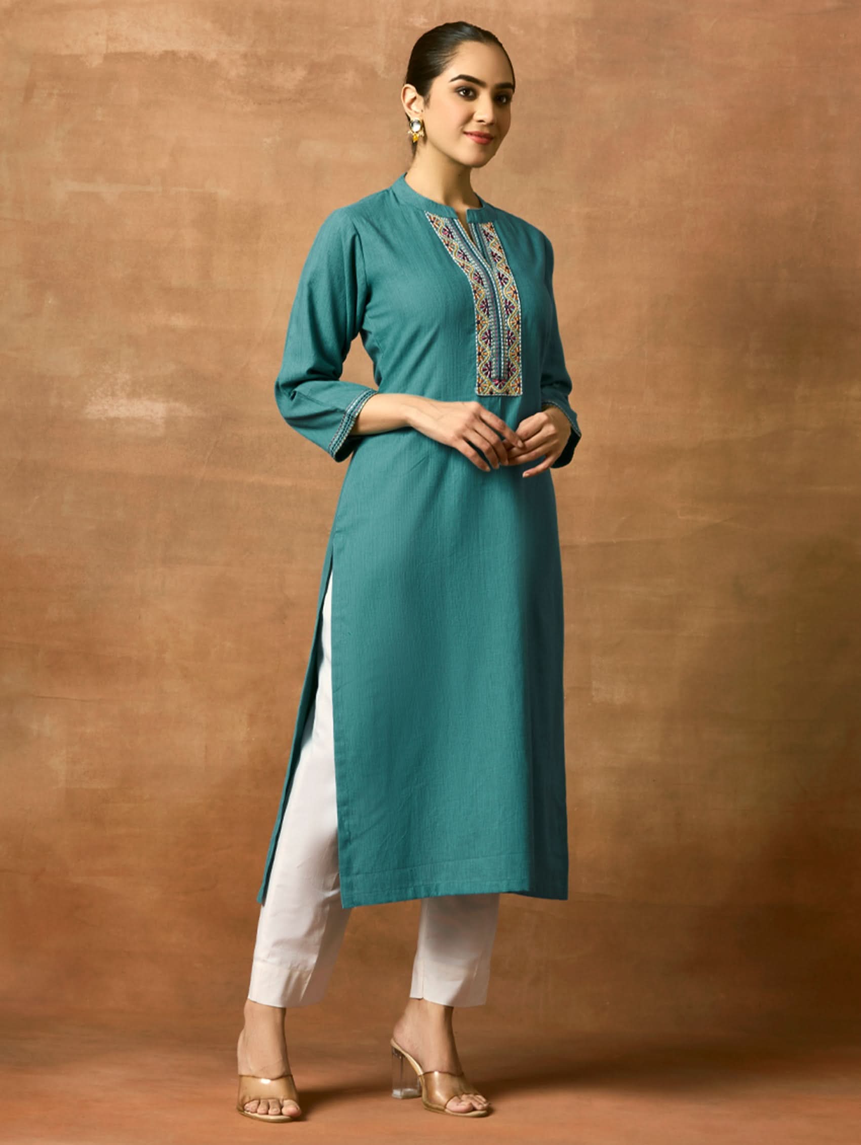Woman wearing a teal kurta with white pants against a brown background