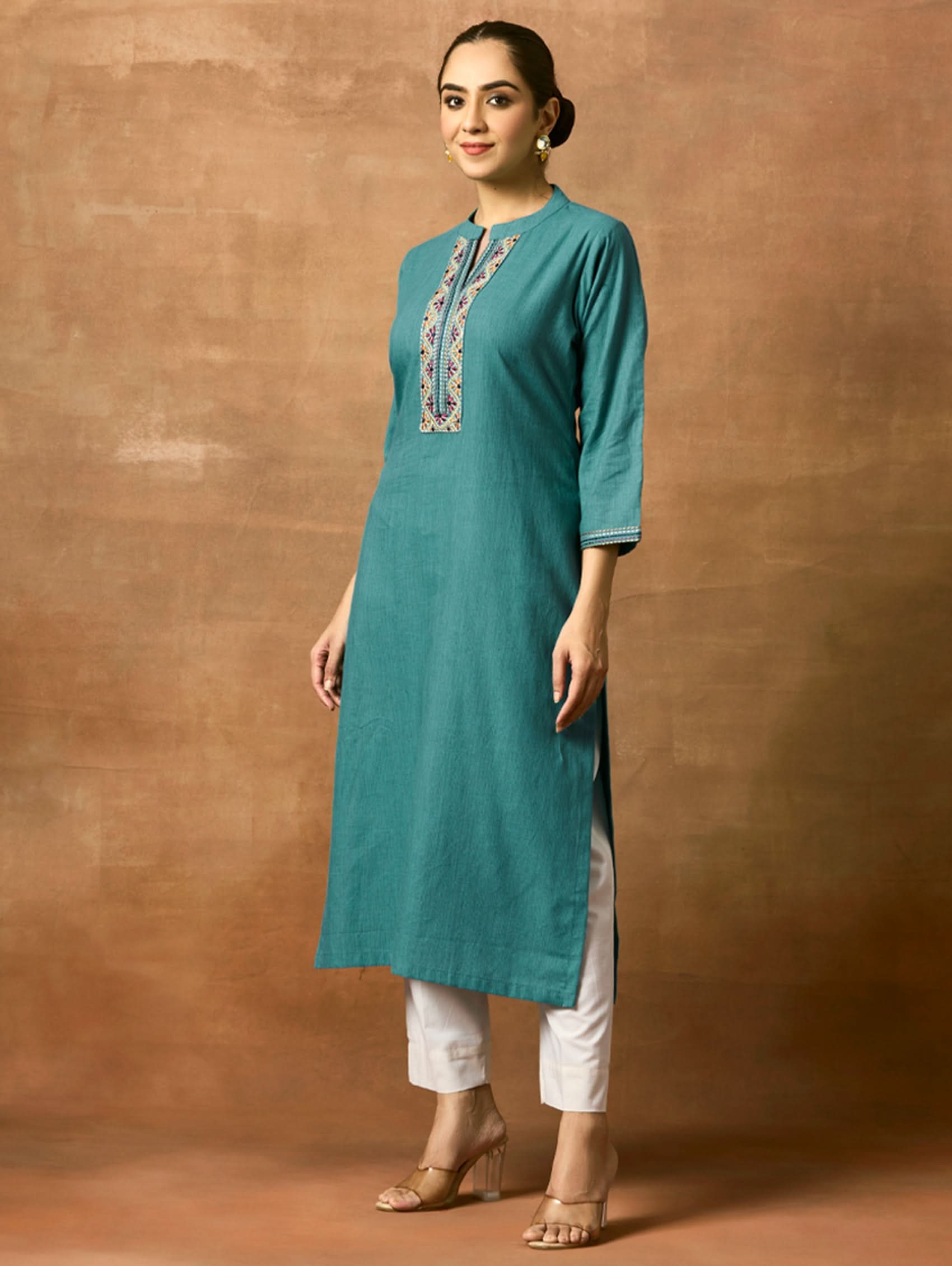 Woman wearing a teal kurta with white pants against a brown background