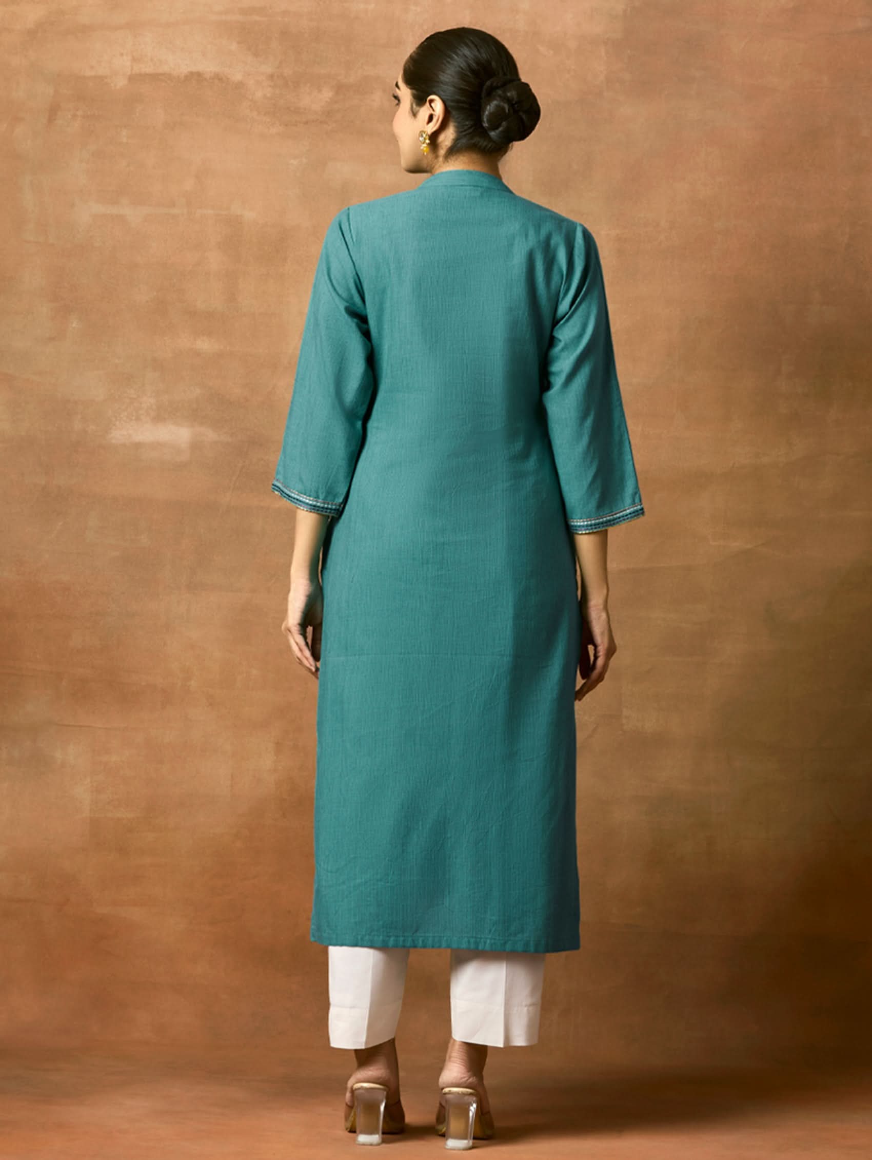 Person wearing a teal kurta and white pants against a brown background