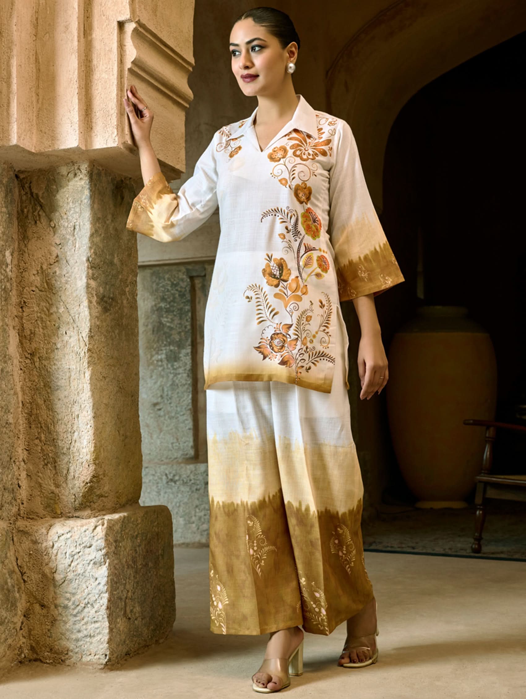 A woman wearing a digitally printed cotton linen short kurta with a revere collar and three-quarter sleeves, paired with straight pants. The outfit features white and golden colors with hand embroidery on the kurta.