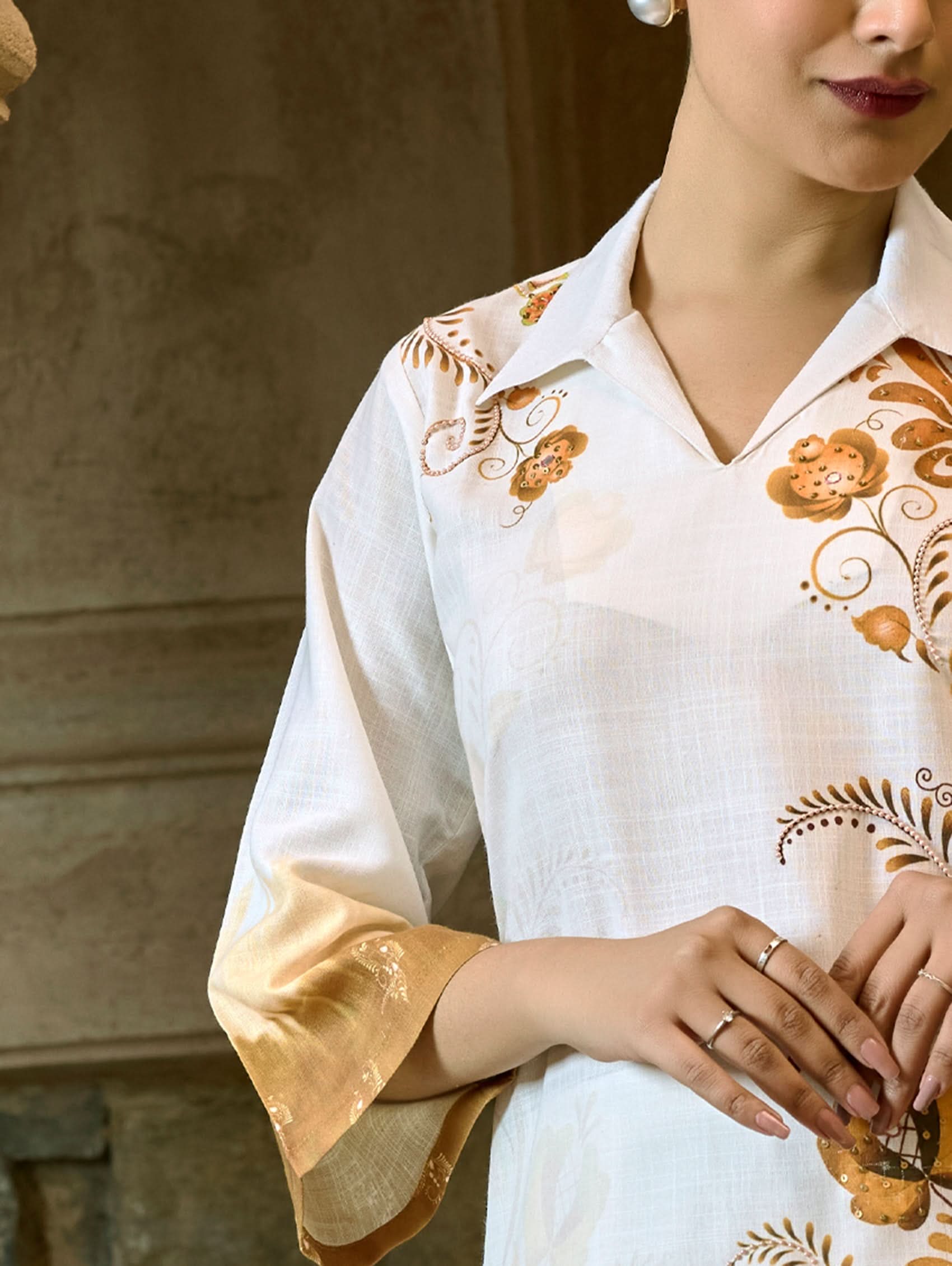 A woman wearing a digitally printed cotton linen short kurta with a revere collar and three-quarter sleeves, paired with straight pants. The outfit features white and golden colors with hand embroidery on the kurta.
